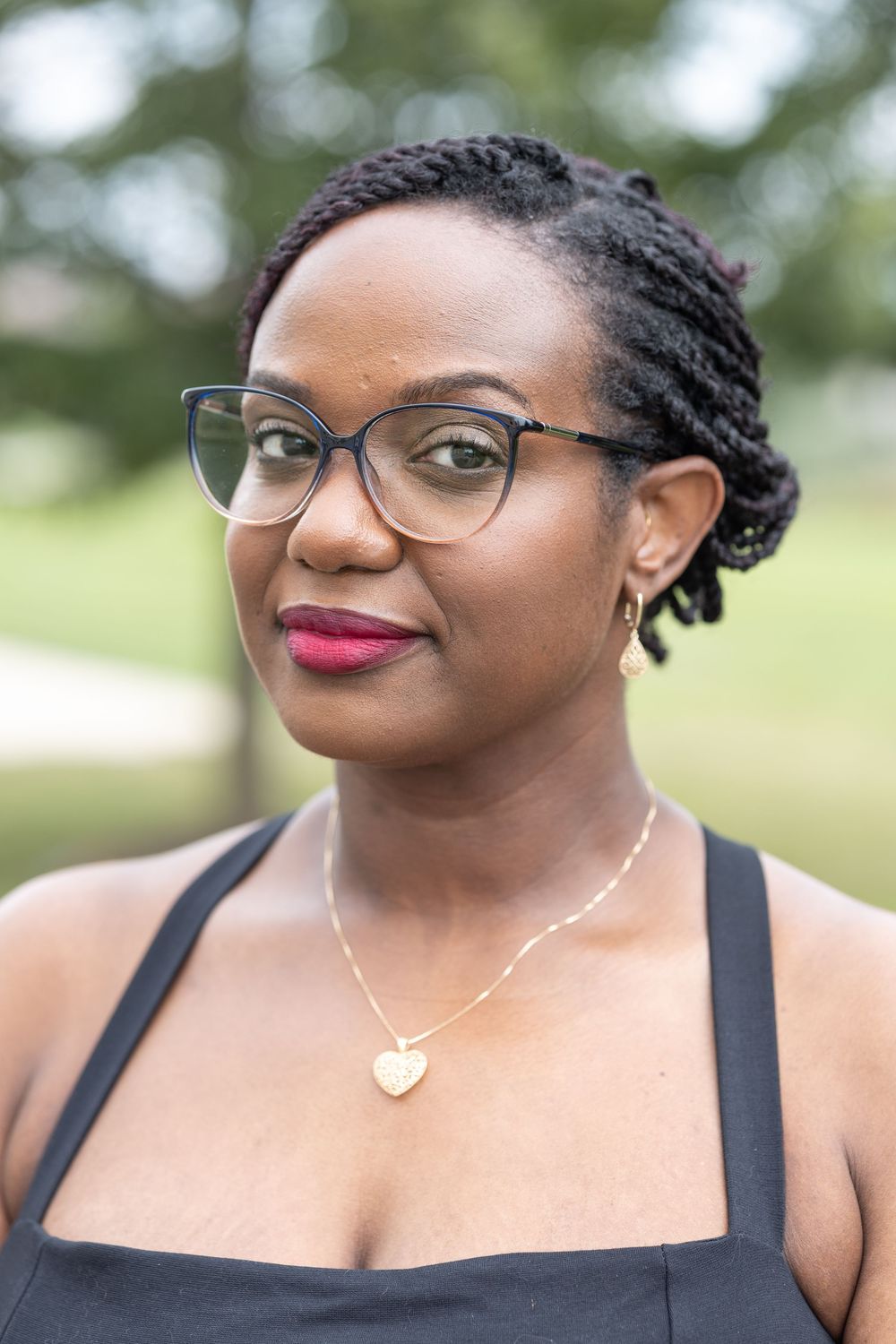 A person wearing glasses and red lipstick smiles warmly at the camera in a black tank top.