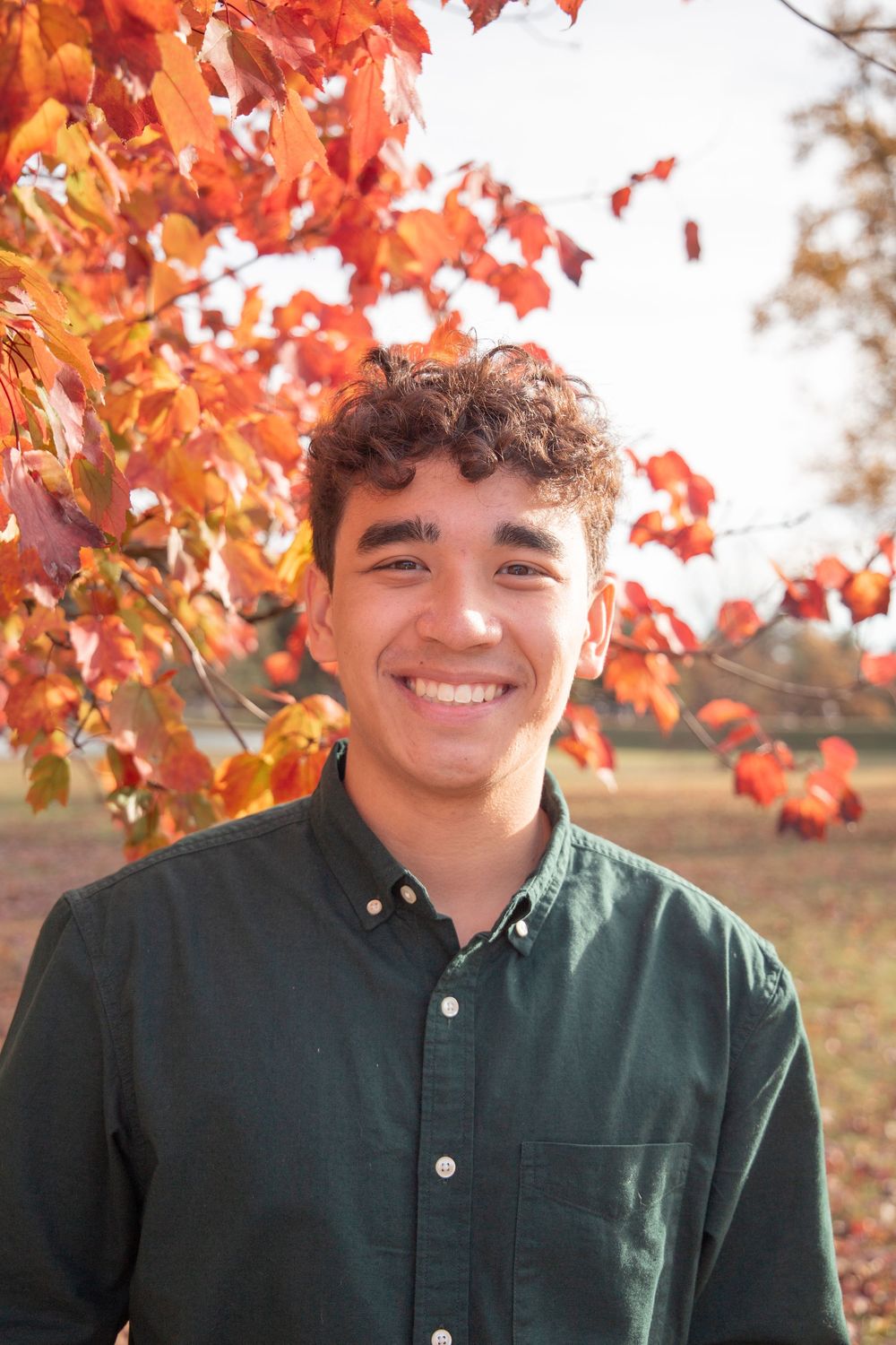 Person wearing a dark green shirt smiles warmly against vibrant orange autumn foliage in outdoor setting.