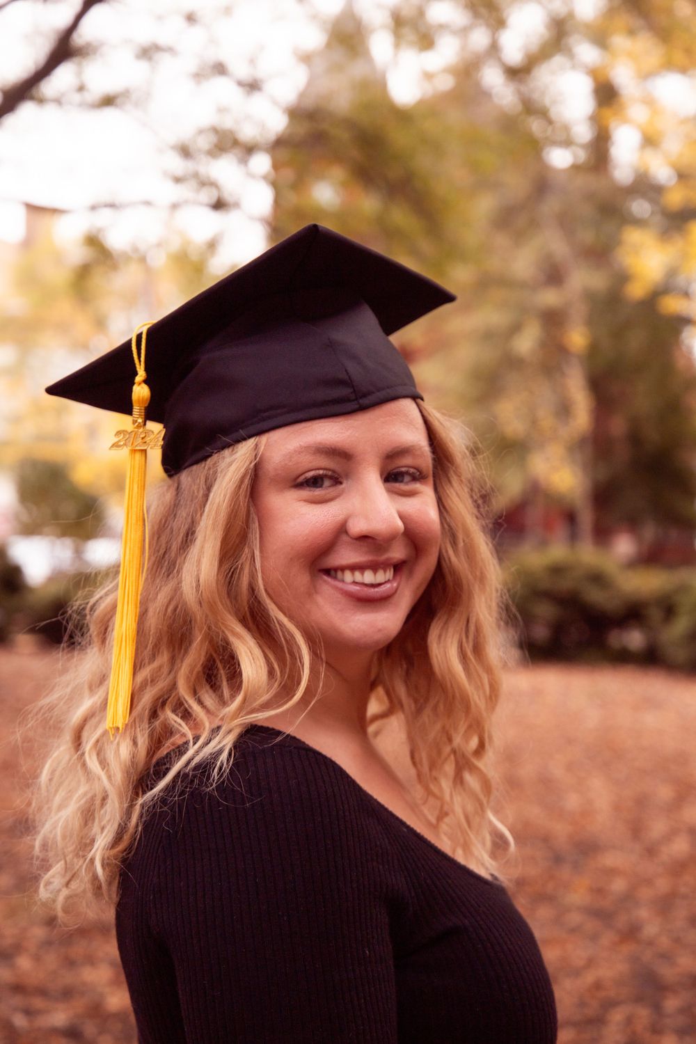 Graduate in black cap and gown with gold tassel poses in autumn outdoor setting.