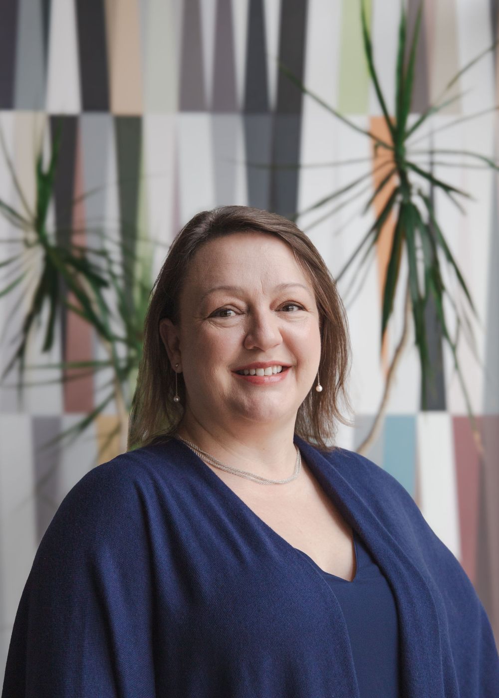Professional indoor headshot with person wearing navy blazer against geometric patterned wall with plants.