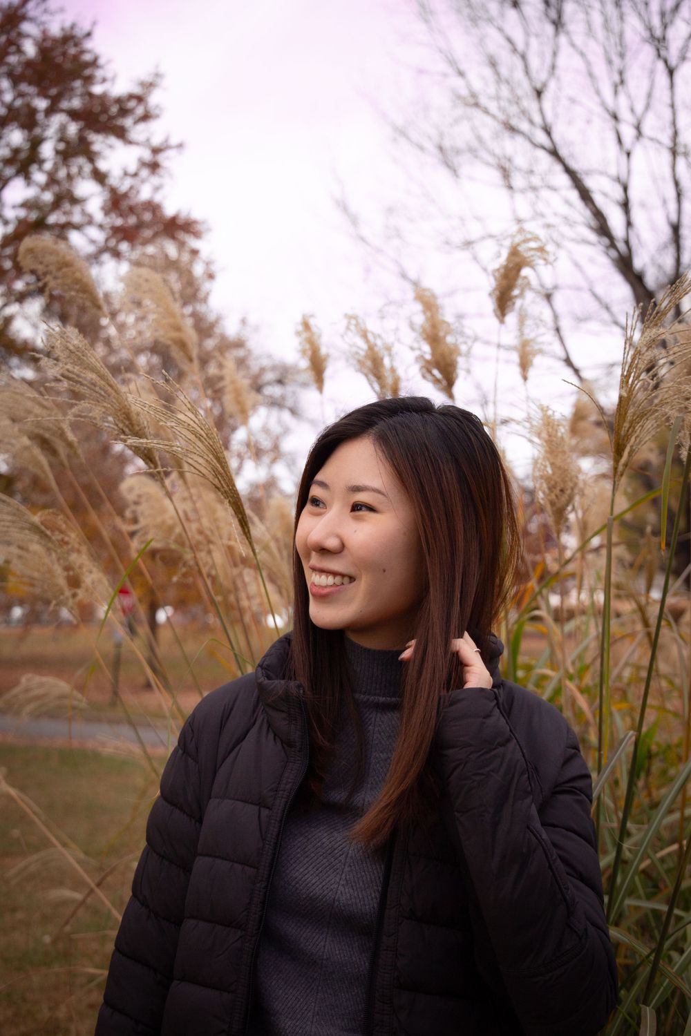 Woman in black puffer jacket smiles while standing among autumn pampas grass.