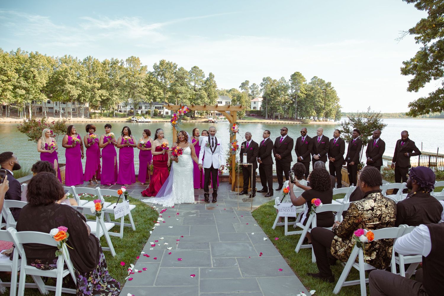 Virginia Wedding Photo & Video Team at Lakeside wedding ceremony with pink-clad bridal party and guests seated on white chairs.
