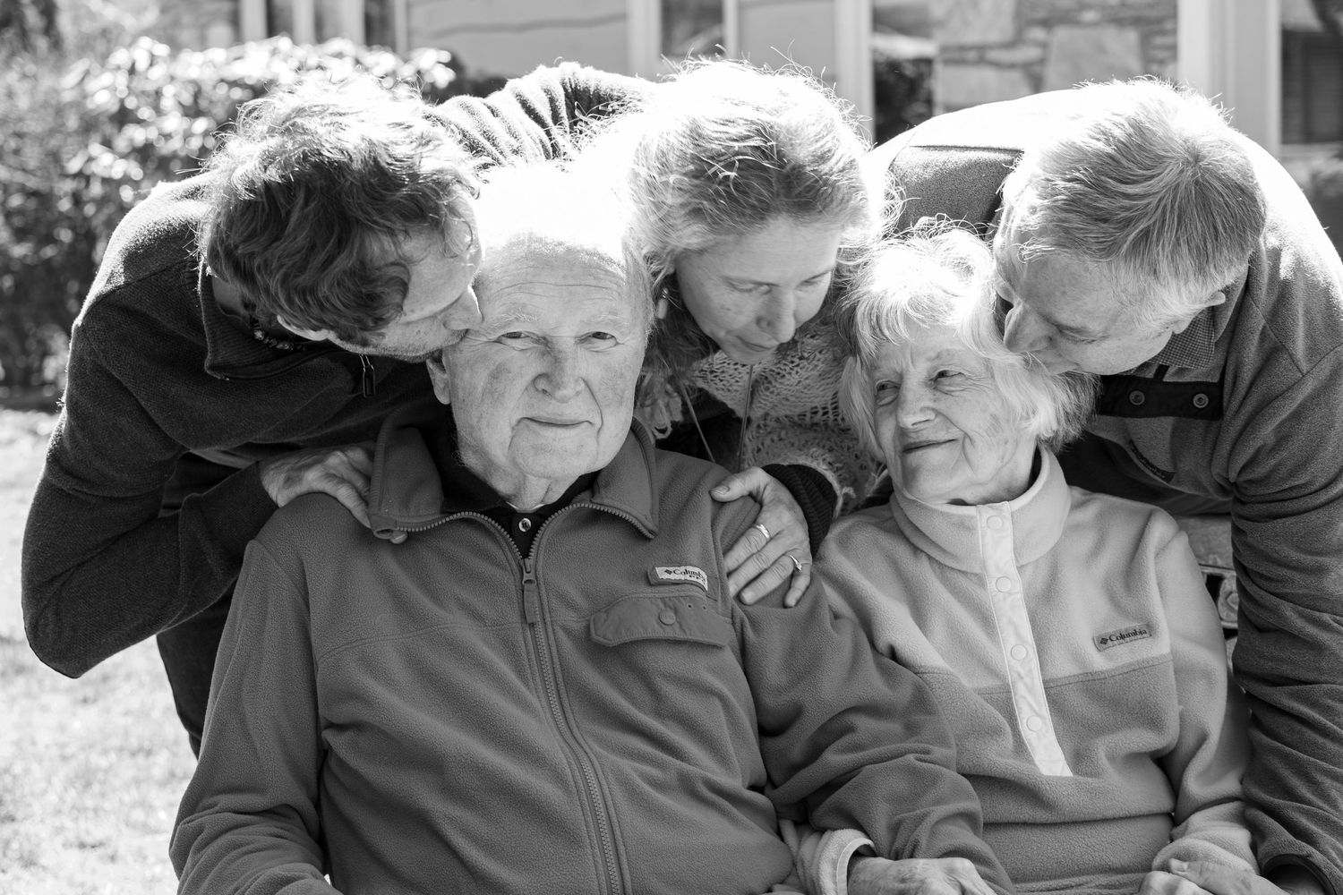 Black and white photo of family members embracing elderly couple outdoors.