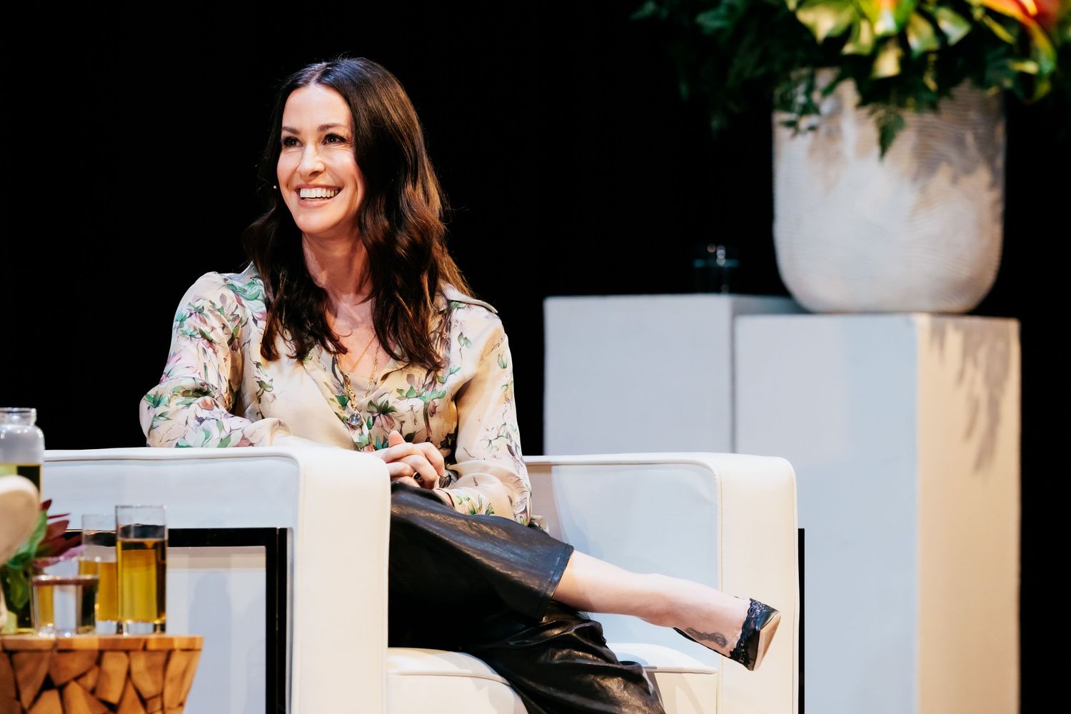 Someone sits comfortably in a white chair during an outdoor conversation, wearing a floral print top and smiling warmly.
