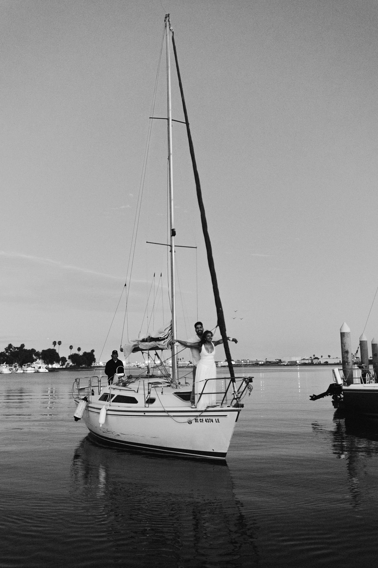 Black and white photo of a sailboat docked in calm marina waters with silhouetted figures on board.