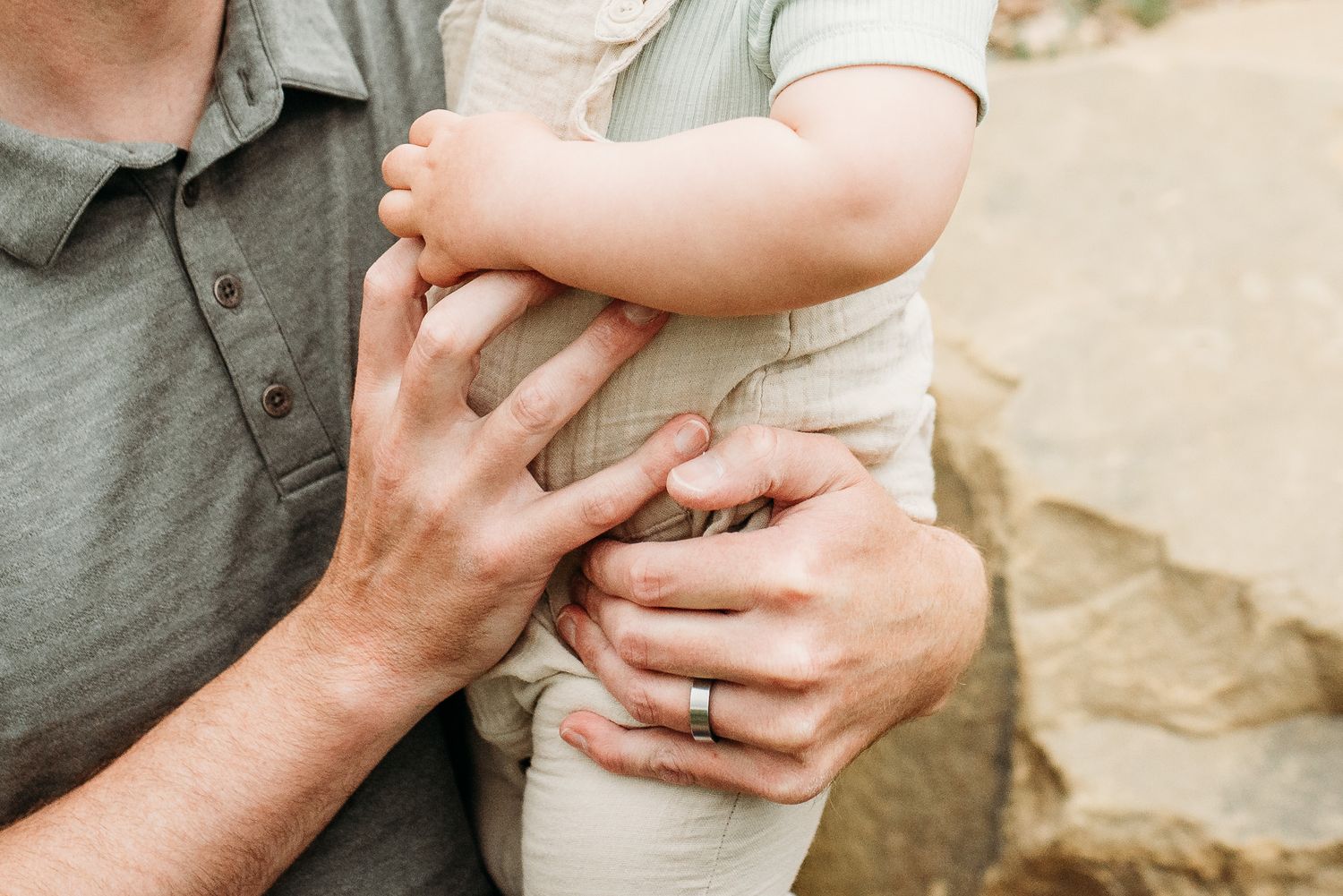 Close-up of adult hands holding a baby wearing a beige outfit, with a wedding ring visible.
