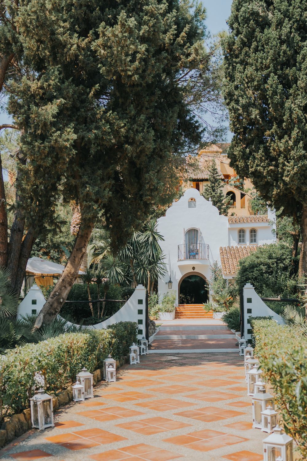 A Spanish-style white stucco venue entrance with terra cotta tiles and lantern-lined path leading to an arched doorway.