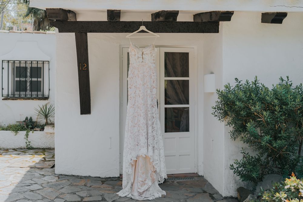 A white wedding dress hangs on display outside a white stucco house with a wooden pergola and green foliage.