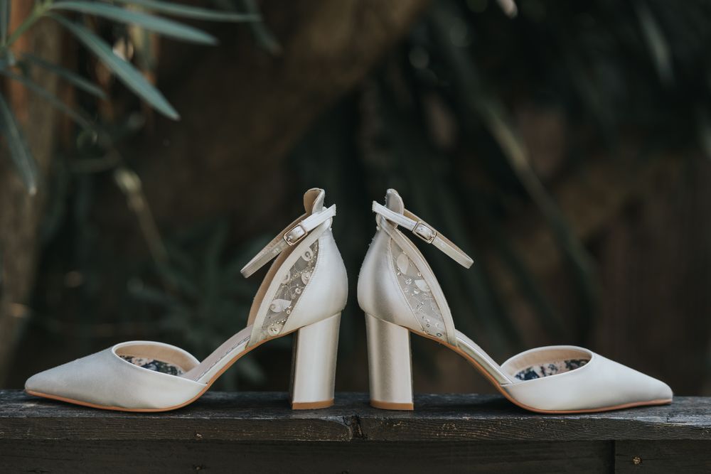 Elegant white bridal shoes with block heels and bow details displayed on a dark wooden surface.