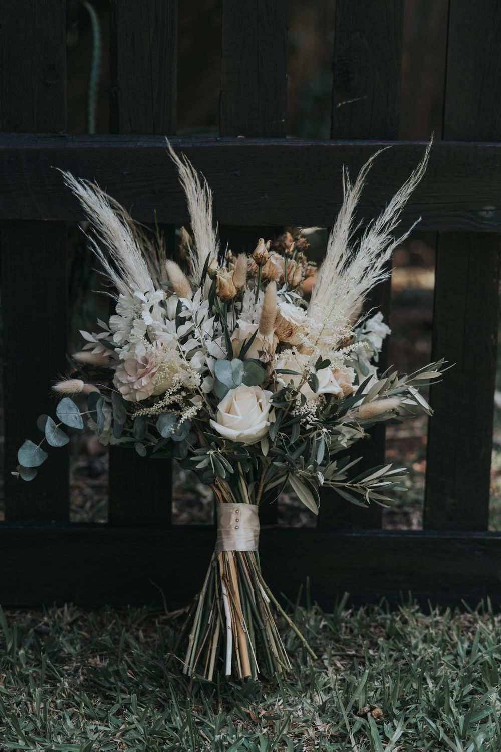 A rustic wedding bouquet with dried pampas grass, white roses, and eucalyptus rests against a dark fence.