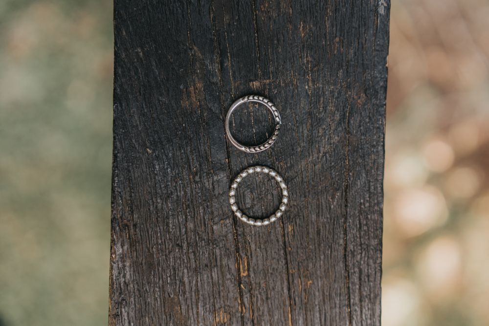 Two silver wedding rings displayed on dark weathered wood with soft bokeh background in natural light.