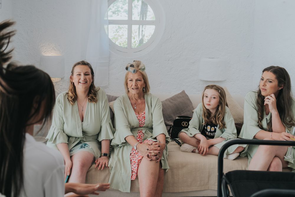 Wedding party relaxes on a couch wearing mint green robes in a bright room with a round window.