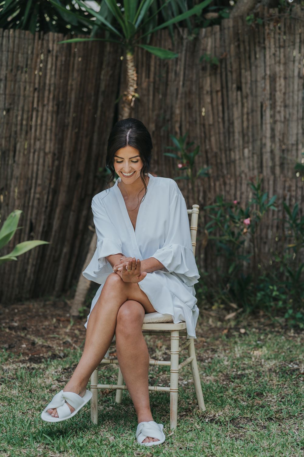 A woman in a white robe sits outdoors on a wooden chair against a bamboo fence wearing casual white slippers.