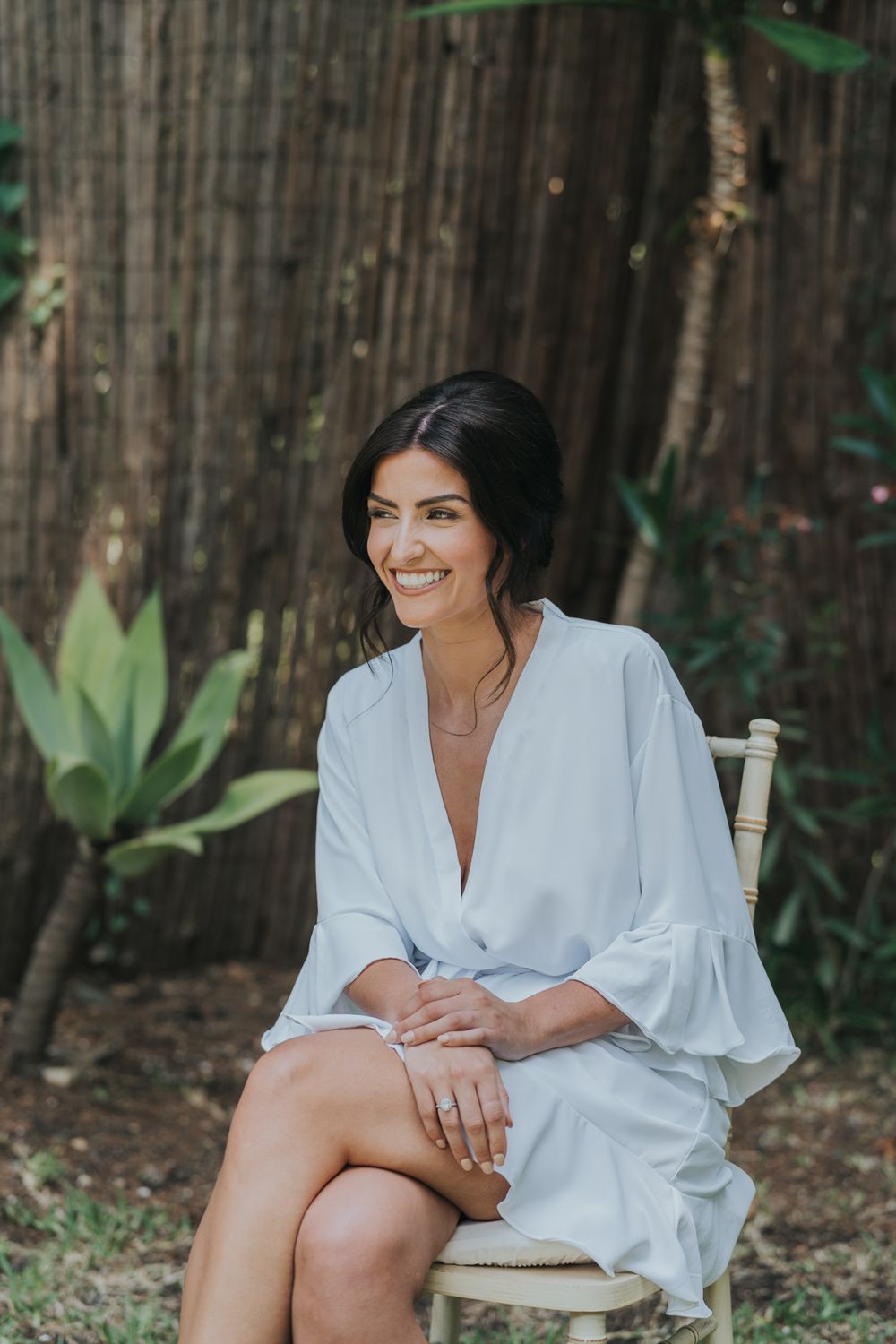 A woman in a white silk robe sits on a chair in a tropical garden setting during wedding preparations.