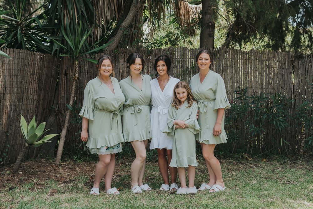 A group wearing sage green dresses poses together in a garden setting with bamboo fencing.