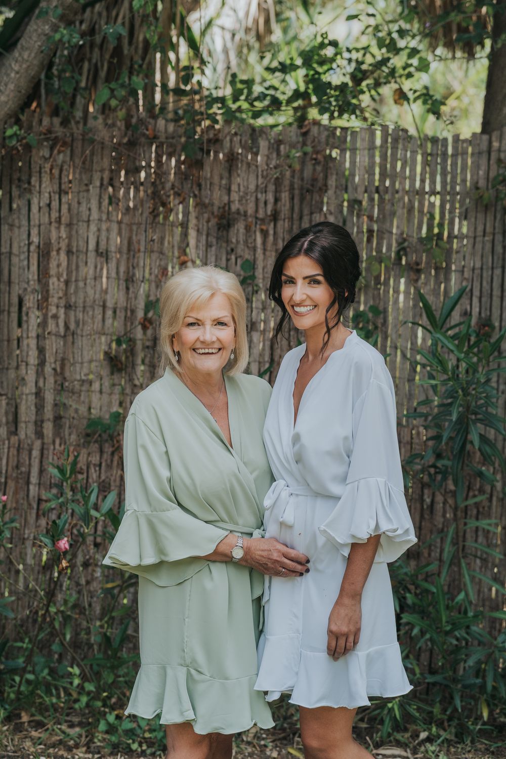 Two women in light-colored dresses pose together against a bamboo fence in a garden setting.