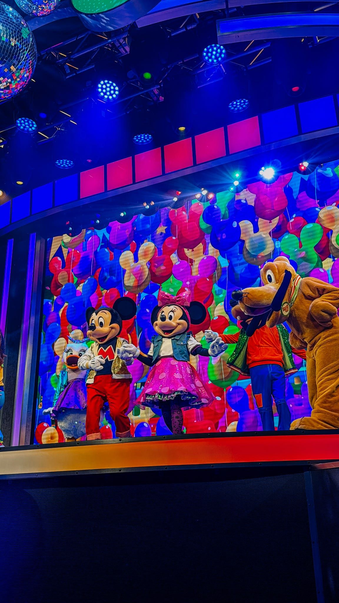 Mickey Mouse, Minnie Mouse, and Pluto Disney characters performing on a colorful stage with balloon decorations.