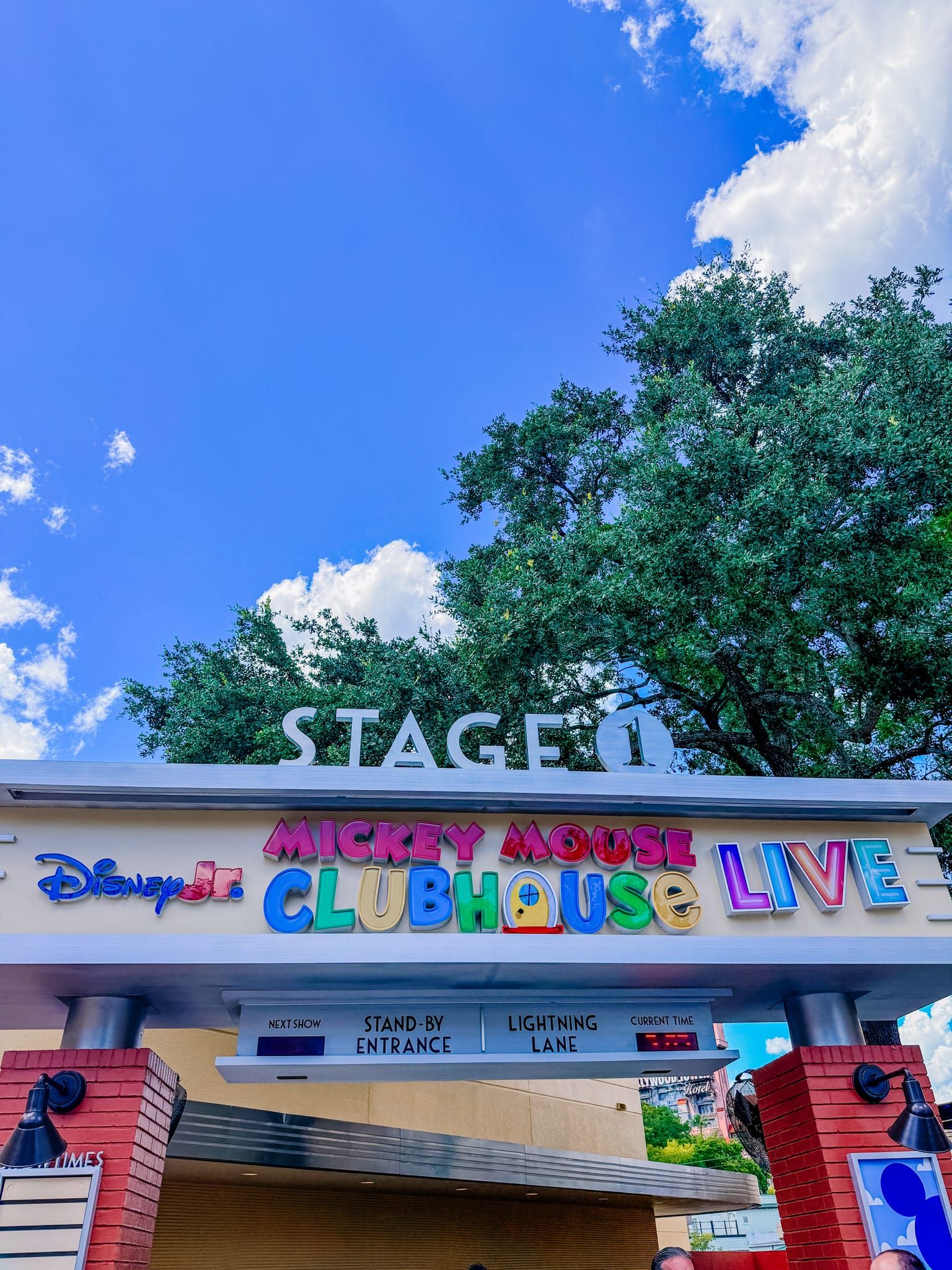 Mickey Mouse Clubhouse Live Stage 1 sign at Disney Jr. with blue sky and trees in background.