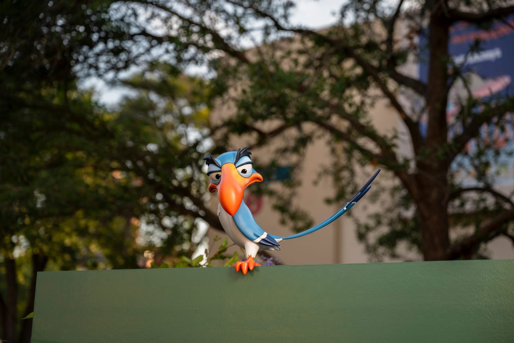 Colorful cartoon bird figurine perched on a green ledge with blurred trees in the background.