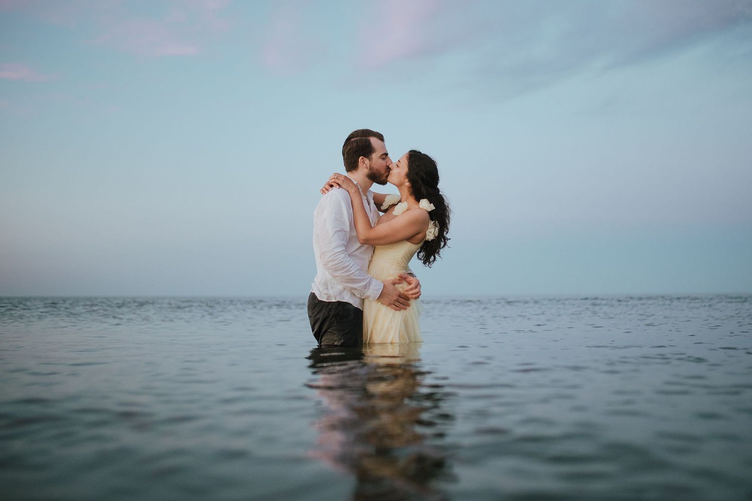 A romantic couple embraces while standing in ocean water at sunset.