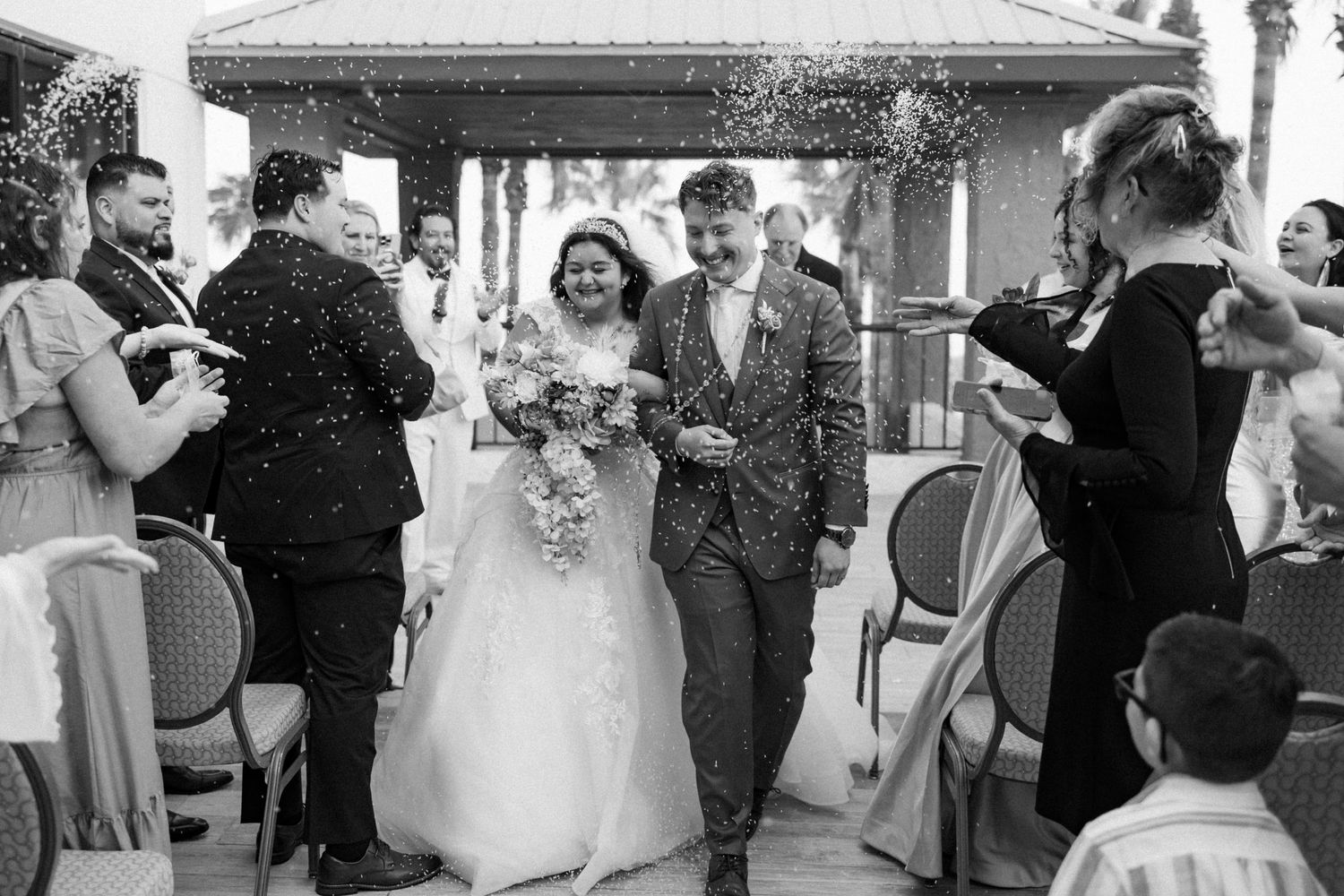 Wedding guests celebrate as newlyweds walk down the aisle at an outdoor ceremony on the beach.