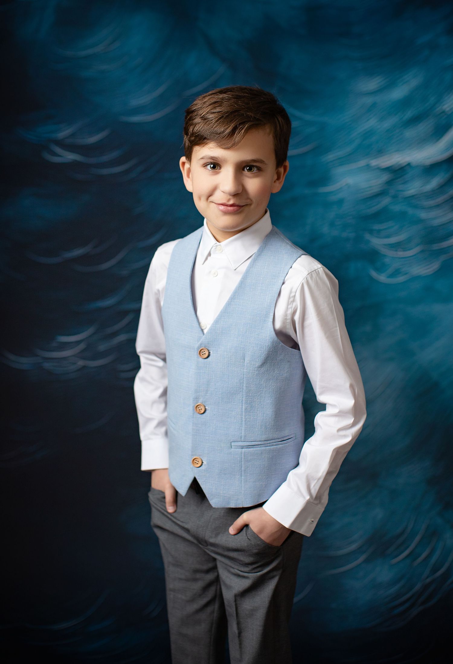 Portrait against a dramatic blue backdrop showing a young student in a light gray vest and white shirt with hands in pockets.
