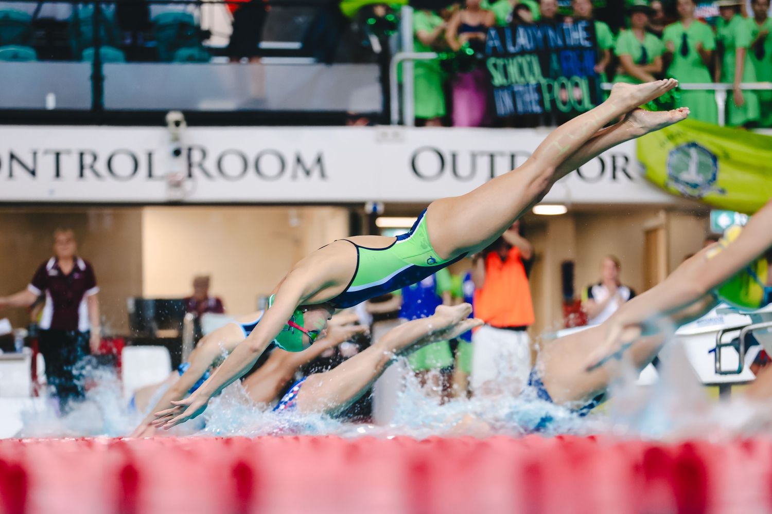 Swimmers diving off starting blocks at swim meet with red lane line and control room visible in background.