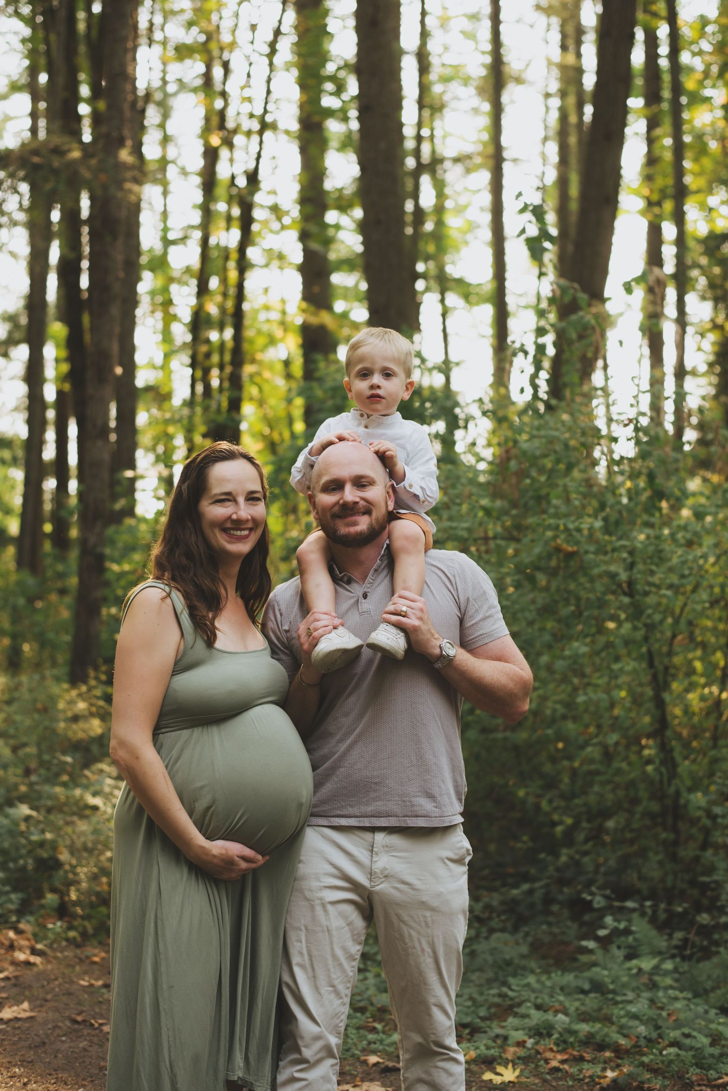 A pregnant woman in a green dress stands with husband and son along a wooded forest trail on a sunny day.