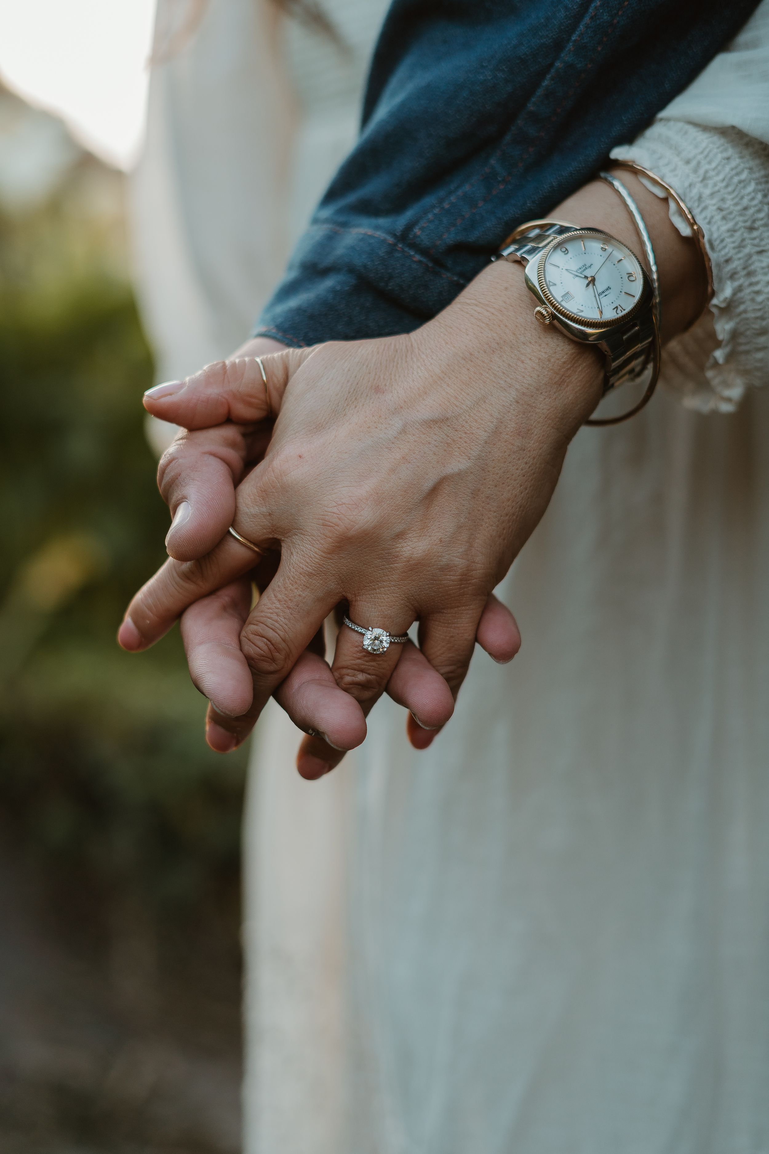 Close-up of hands holding each other with engagement ring and wristwatch visible on a blurred outdoor background.