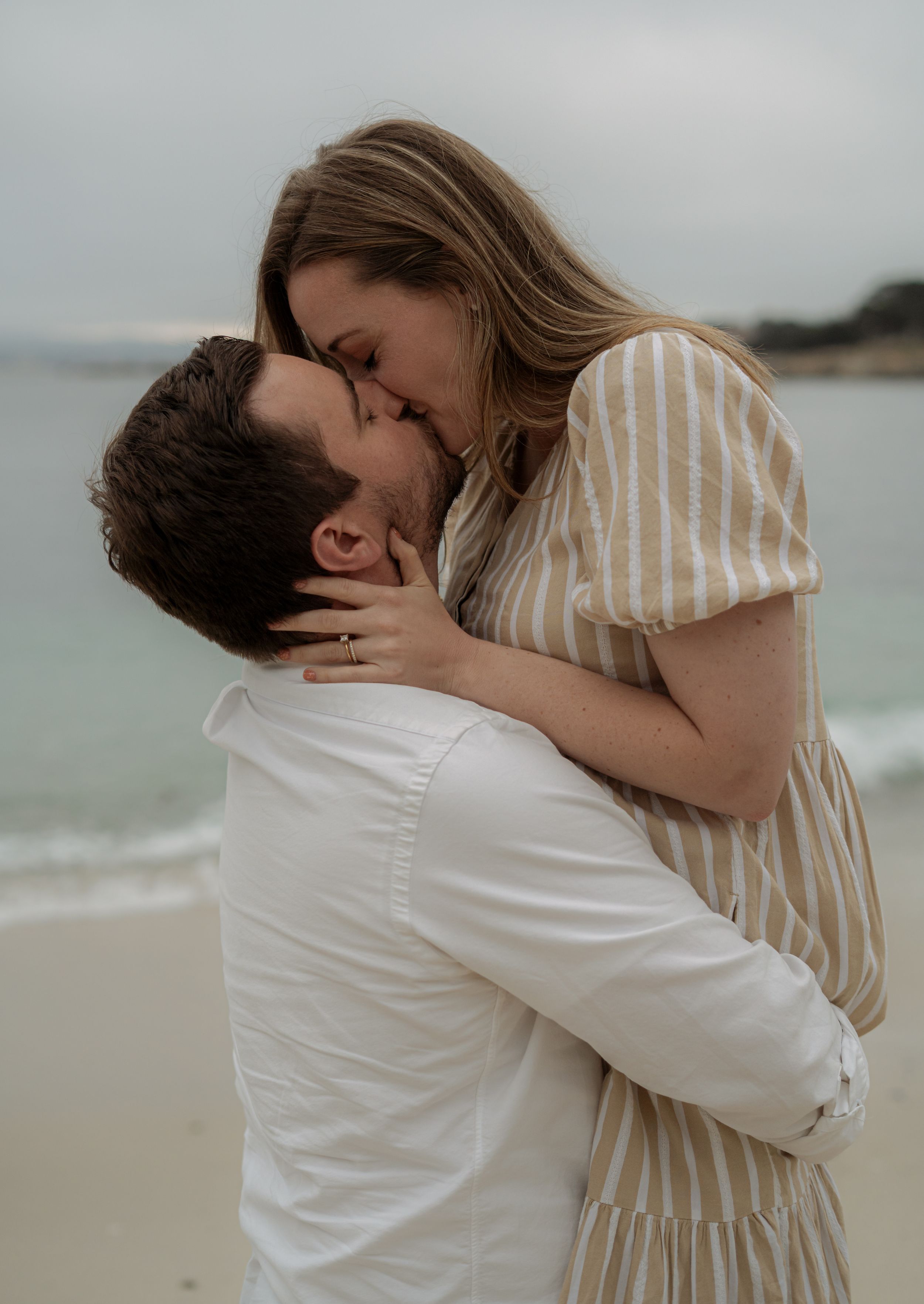 A romantic couple shares an intimate embrace on a cloudy beach, with the woman in a striped dress and man in white shirt.