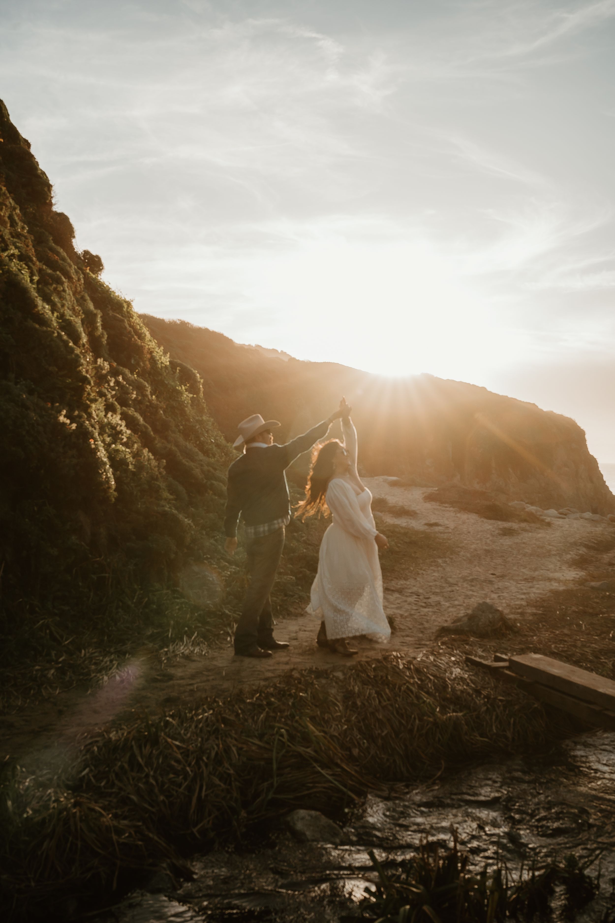 Two figures dance together on a dramatic coastal cliff at sunset, with golden sunlight streaming through the mountainous landscape.