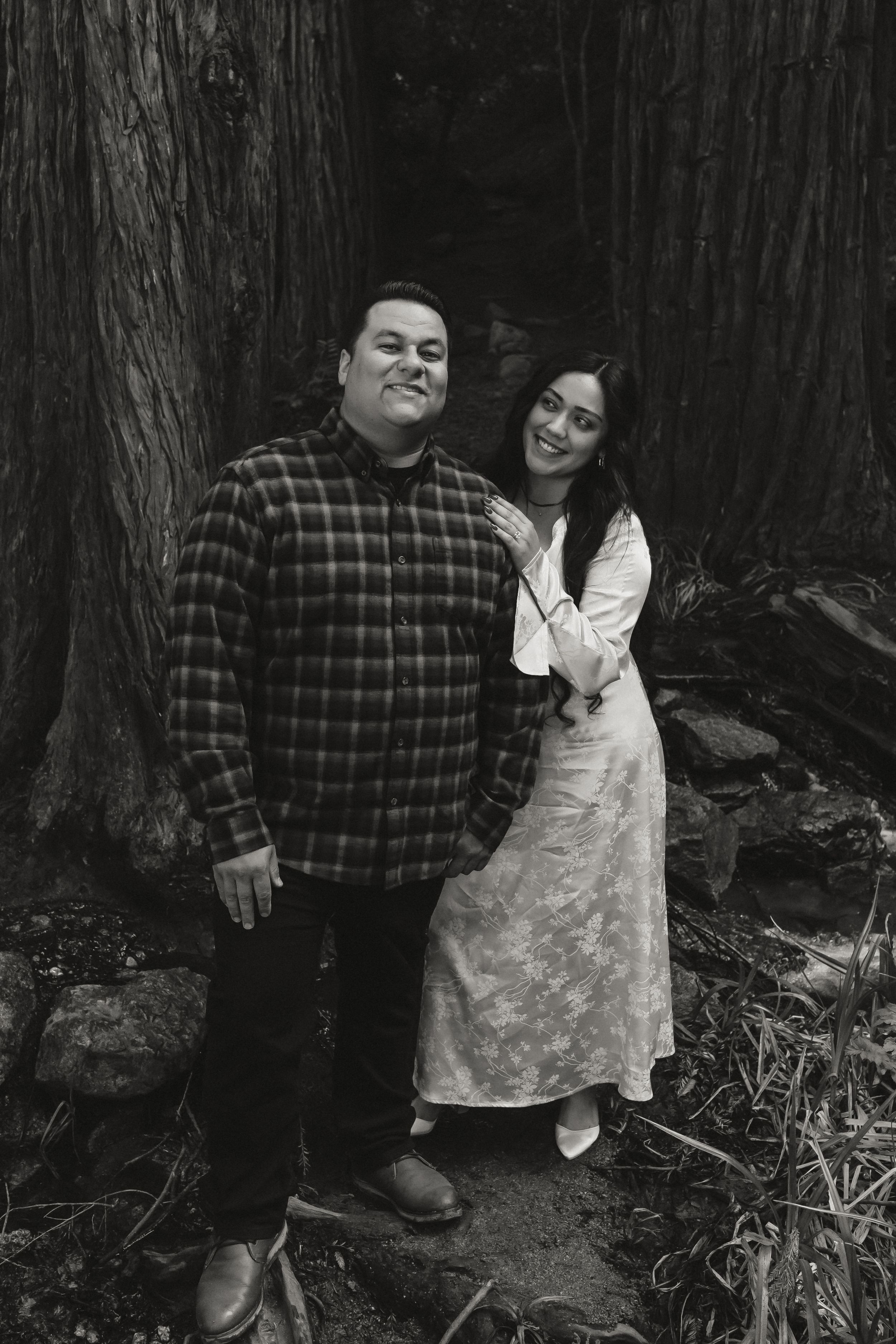 A black and white portrait of a couple standing in front of large tree trunks in a forest setting.