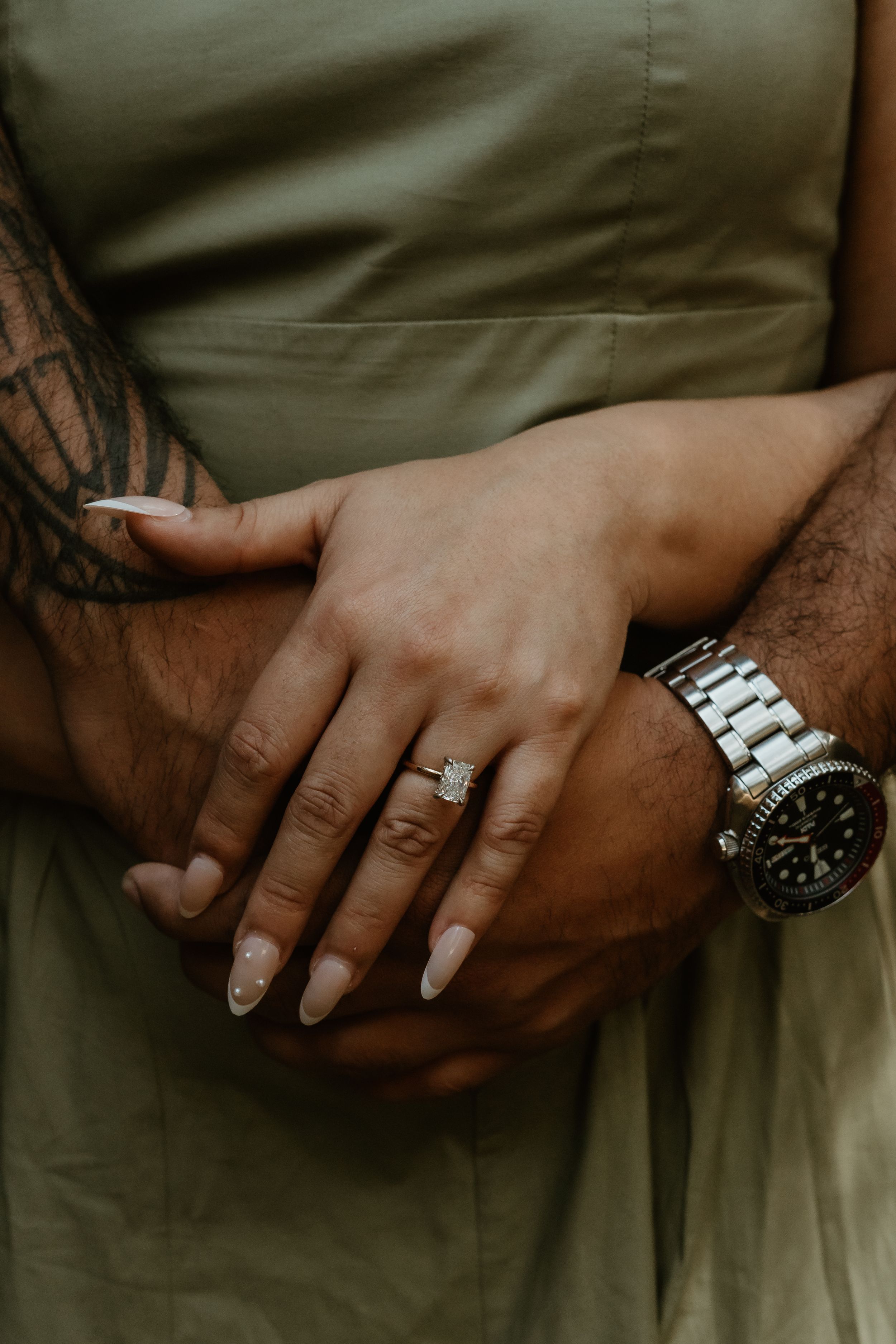 Close-up of hands with engagement ring, silver watch, and tattoo resting against an olive green fabric background.