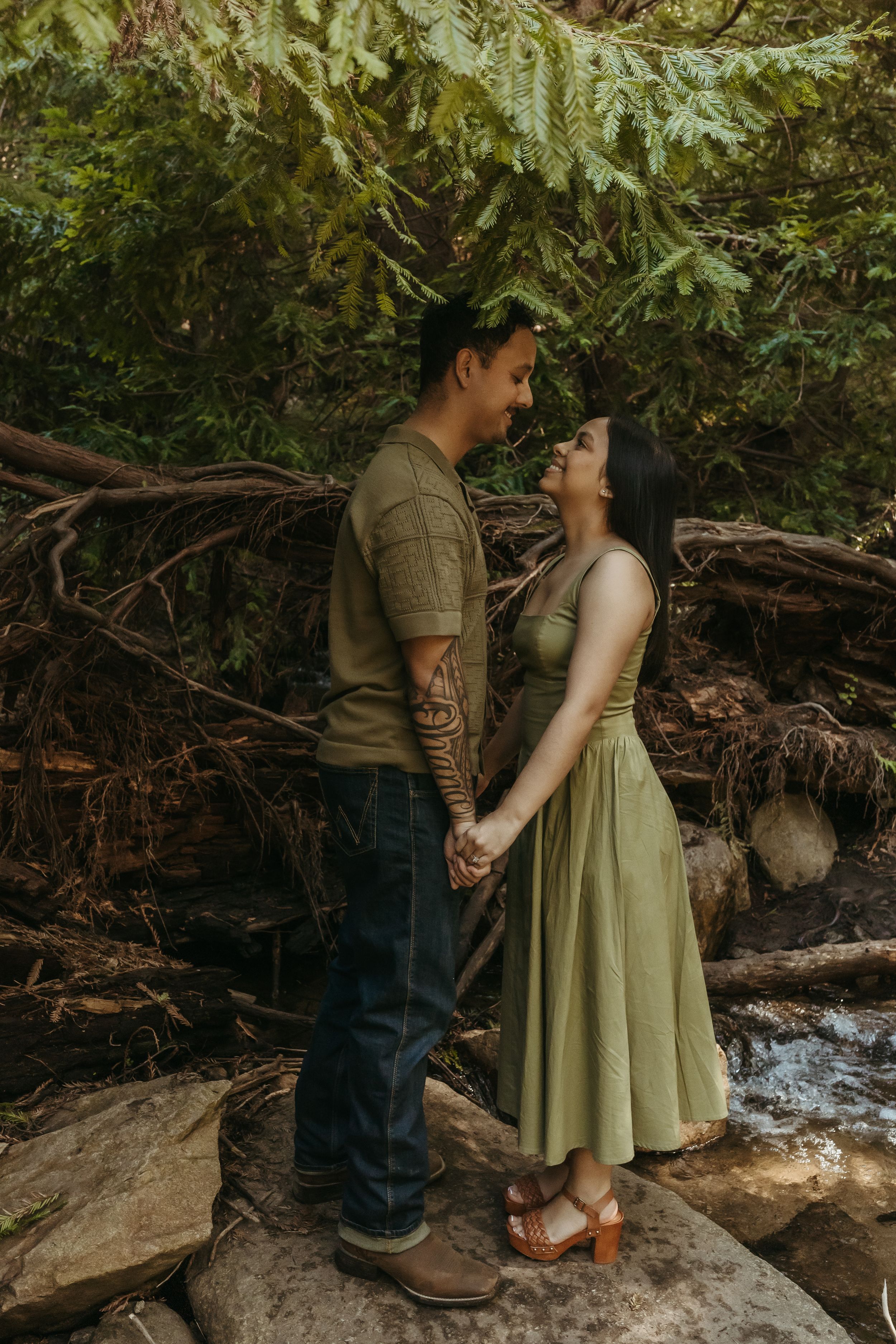 A romantic couple stands together on rocks by a stream in a dreamy forest setting with green foliage overhead.