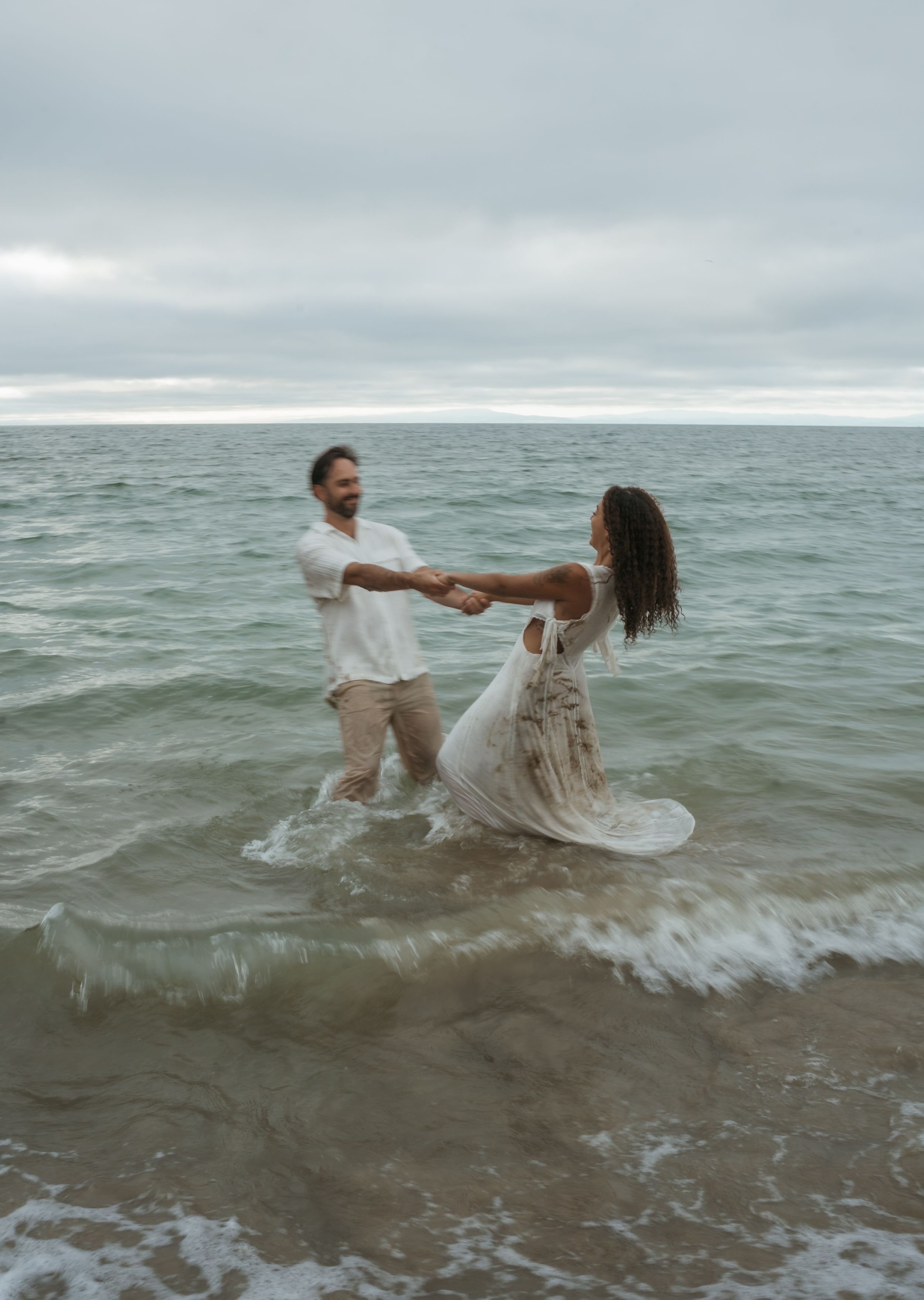 A couple wearing white clothing plays in ocean waves during an overcast day.