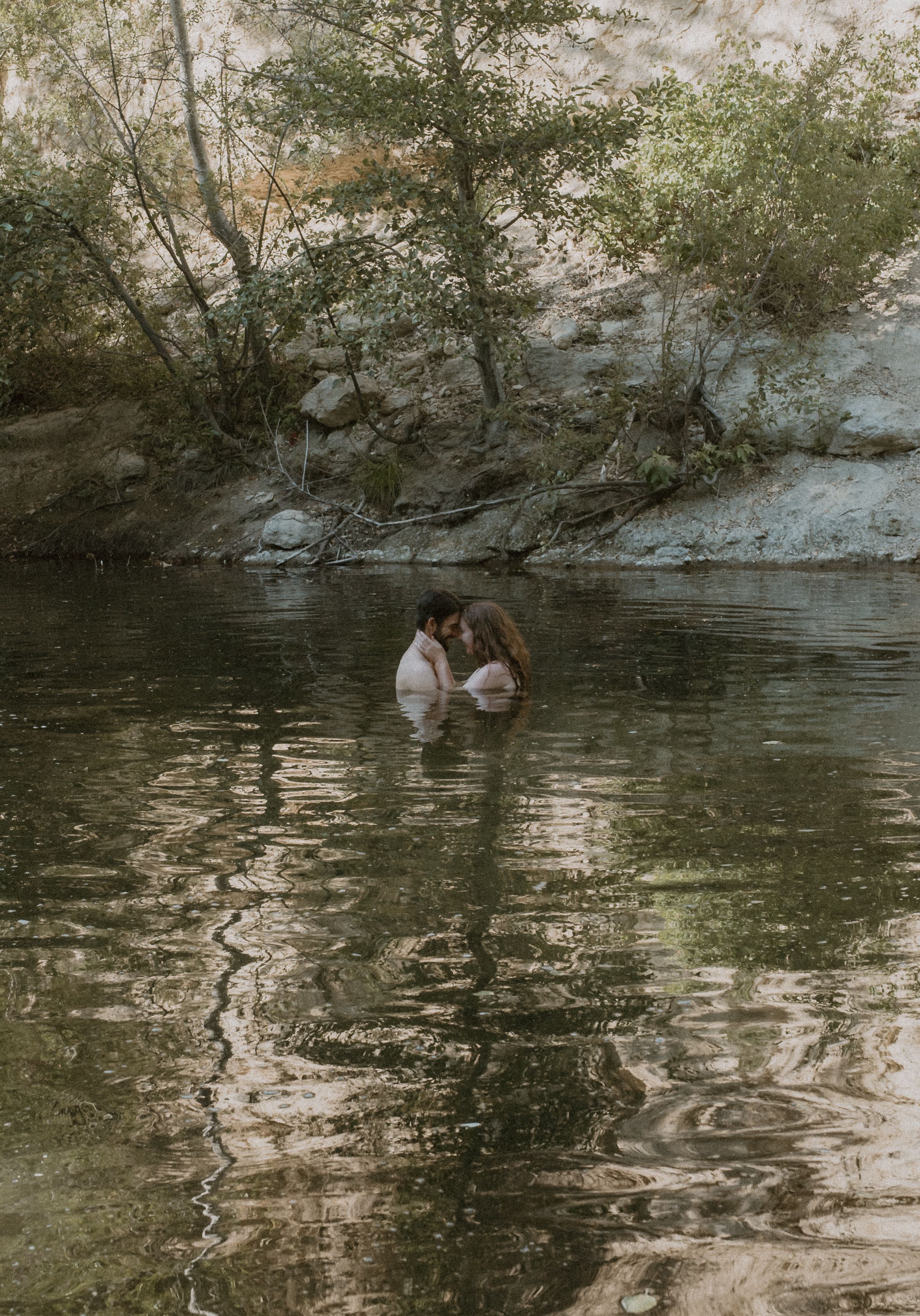 Two silhouettes stand in a tranquil forest pond with reflective water and overhanging trees.