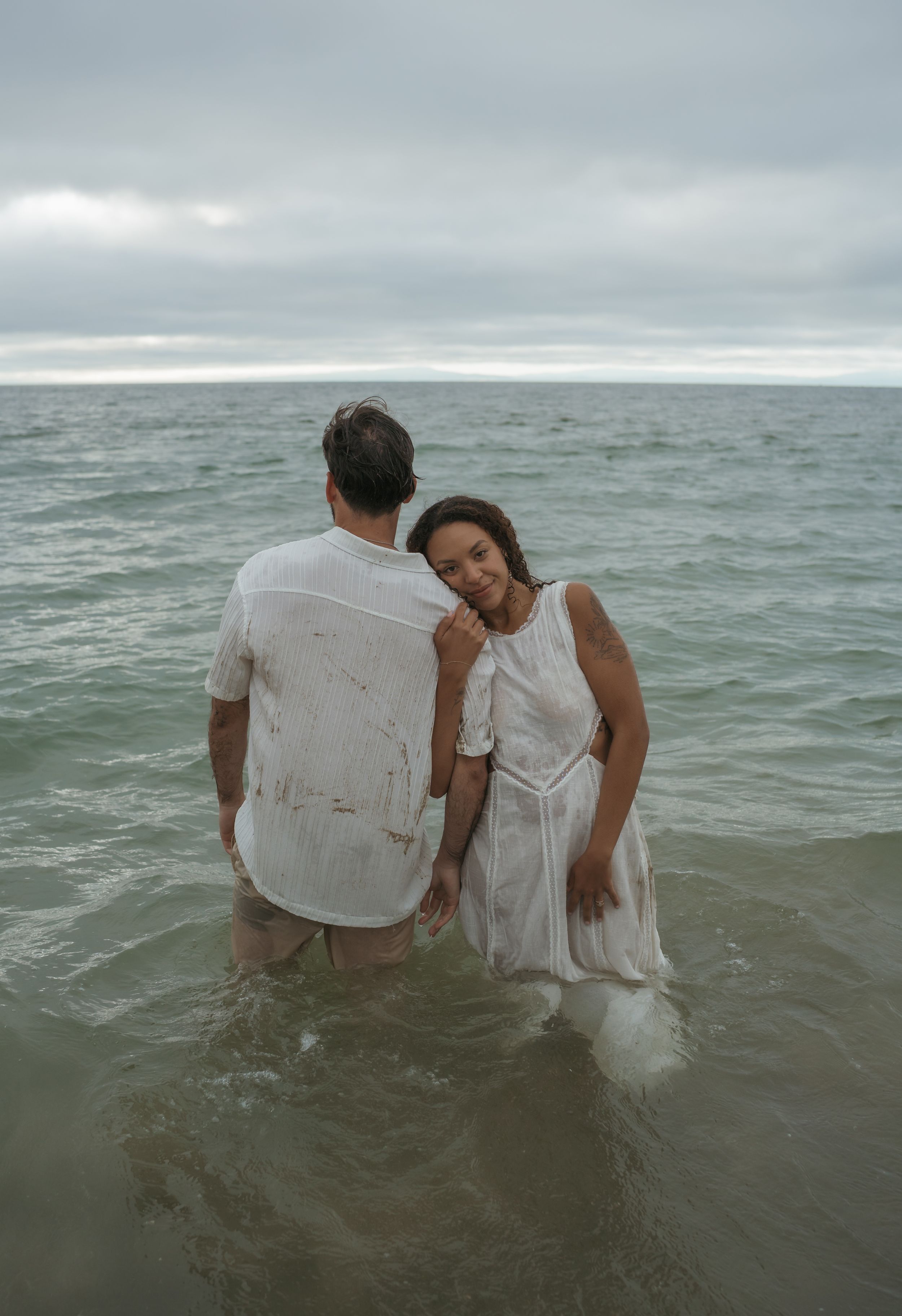 A couple sits together in the ocean waves wearing white clothes in a series of romantic beach photos.