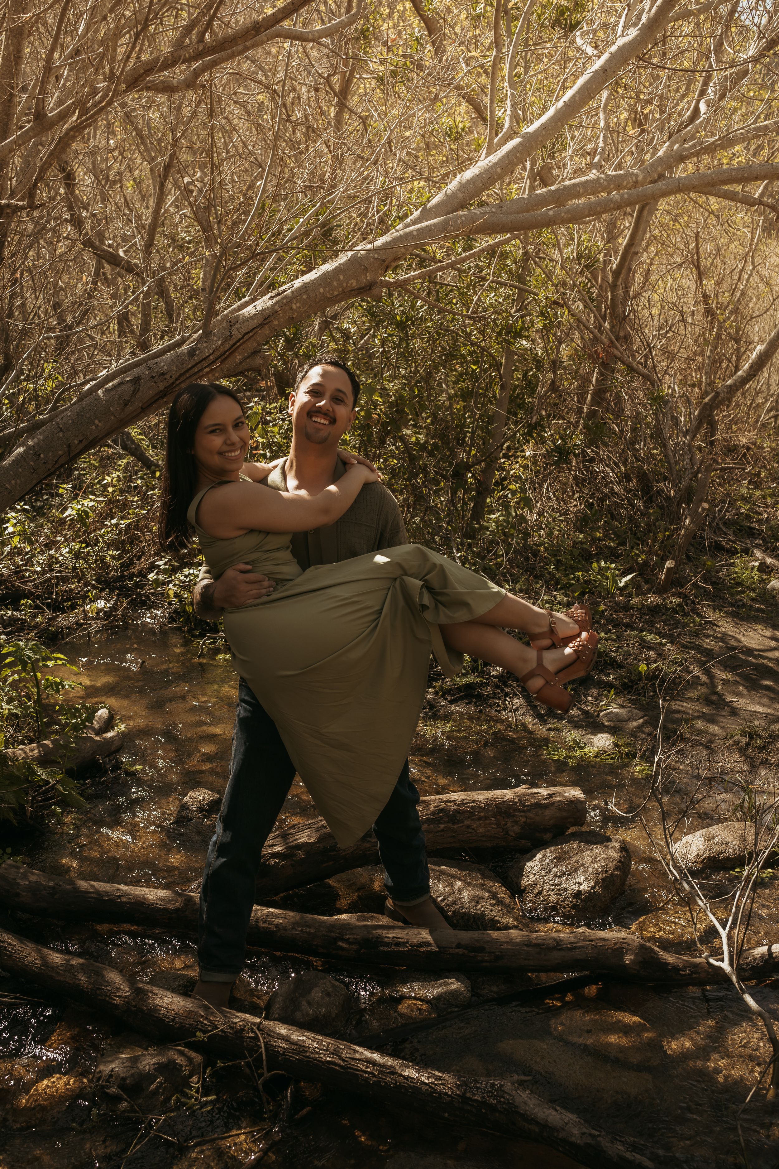 A couple share a romantic moment outdoors in a wooded area with dappled sunlight filtering through the branches.