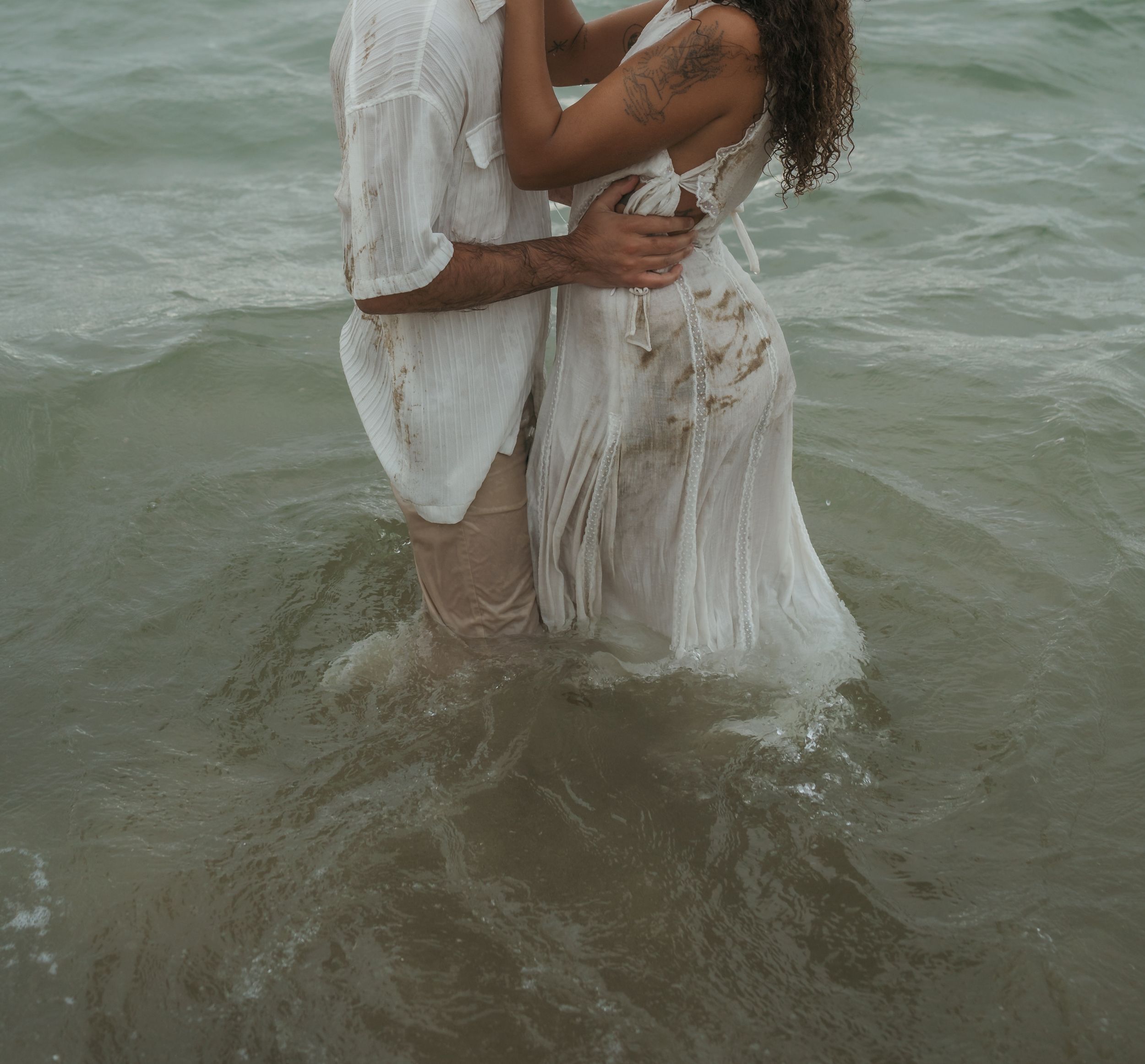 A couple embraces while standing in shallow ocean waters, wearing wet white clothing.