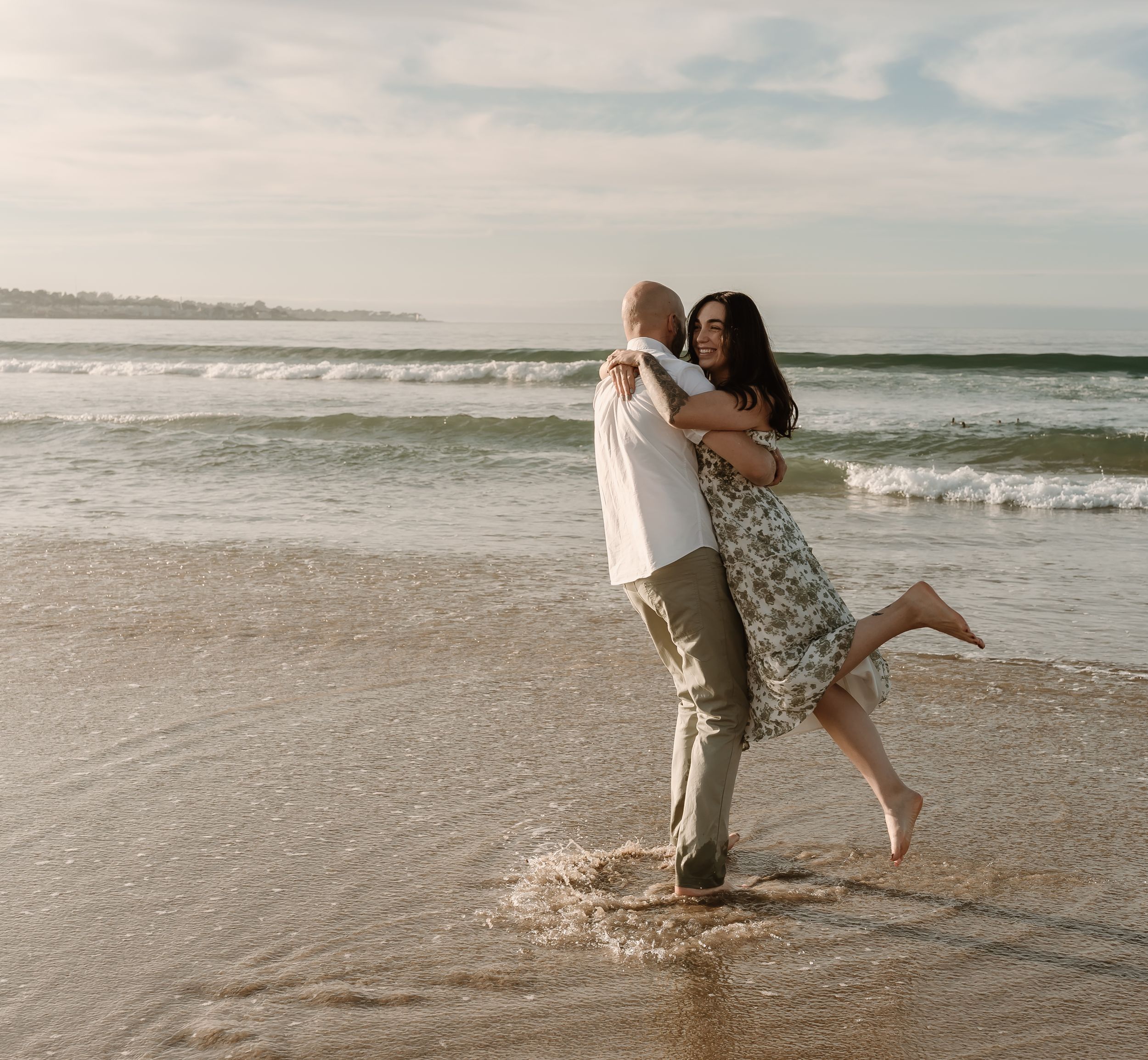 A couple embraces while standing in the ocean surf at sunset on a sandy beach.