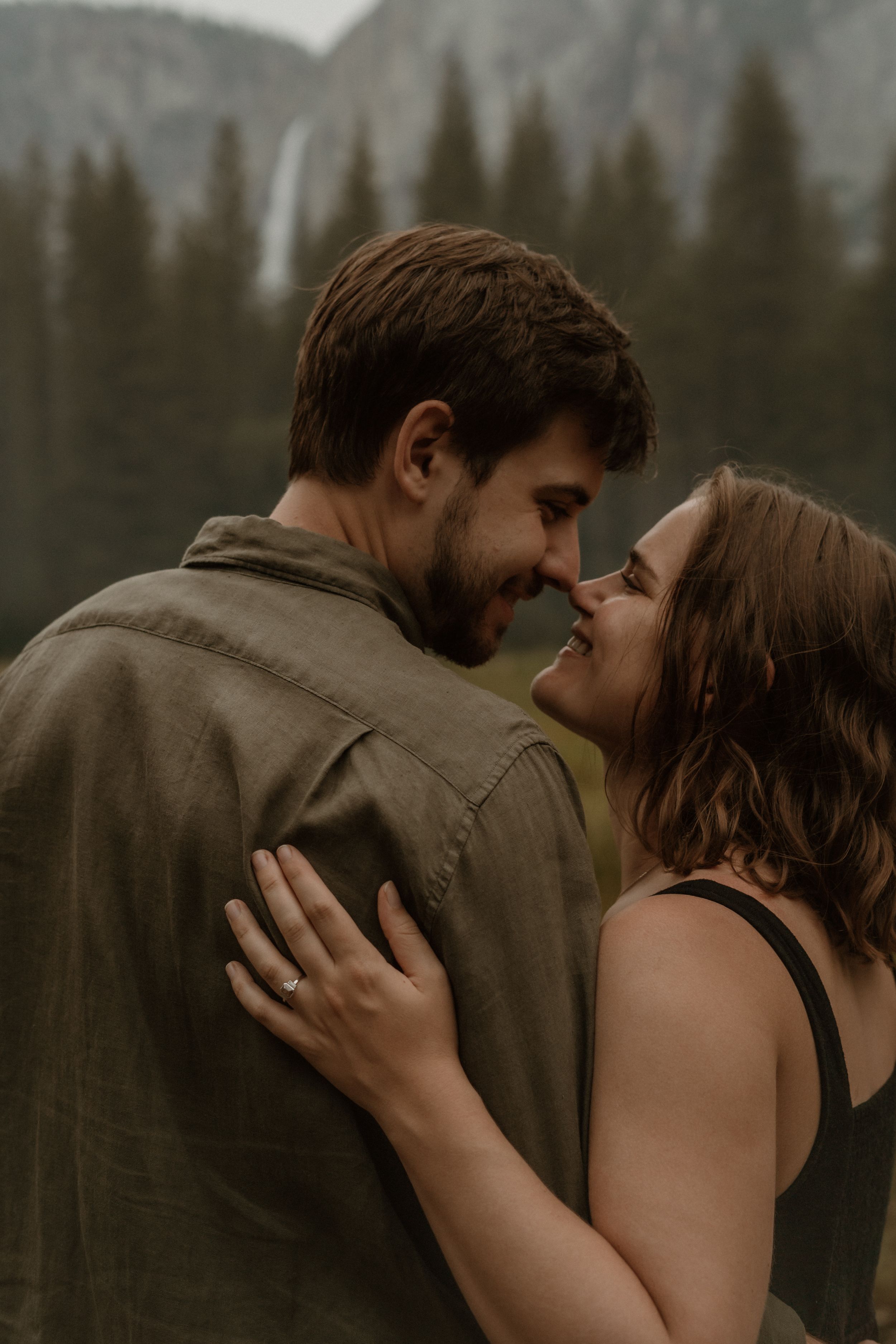 A couple in brown clothing share an intimate embrace against a moody forest backdrop.