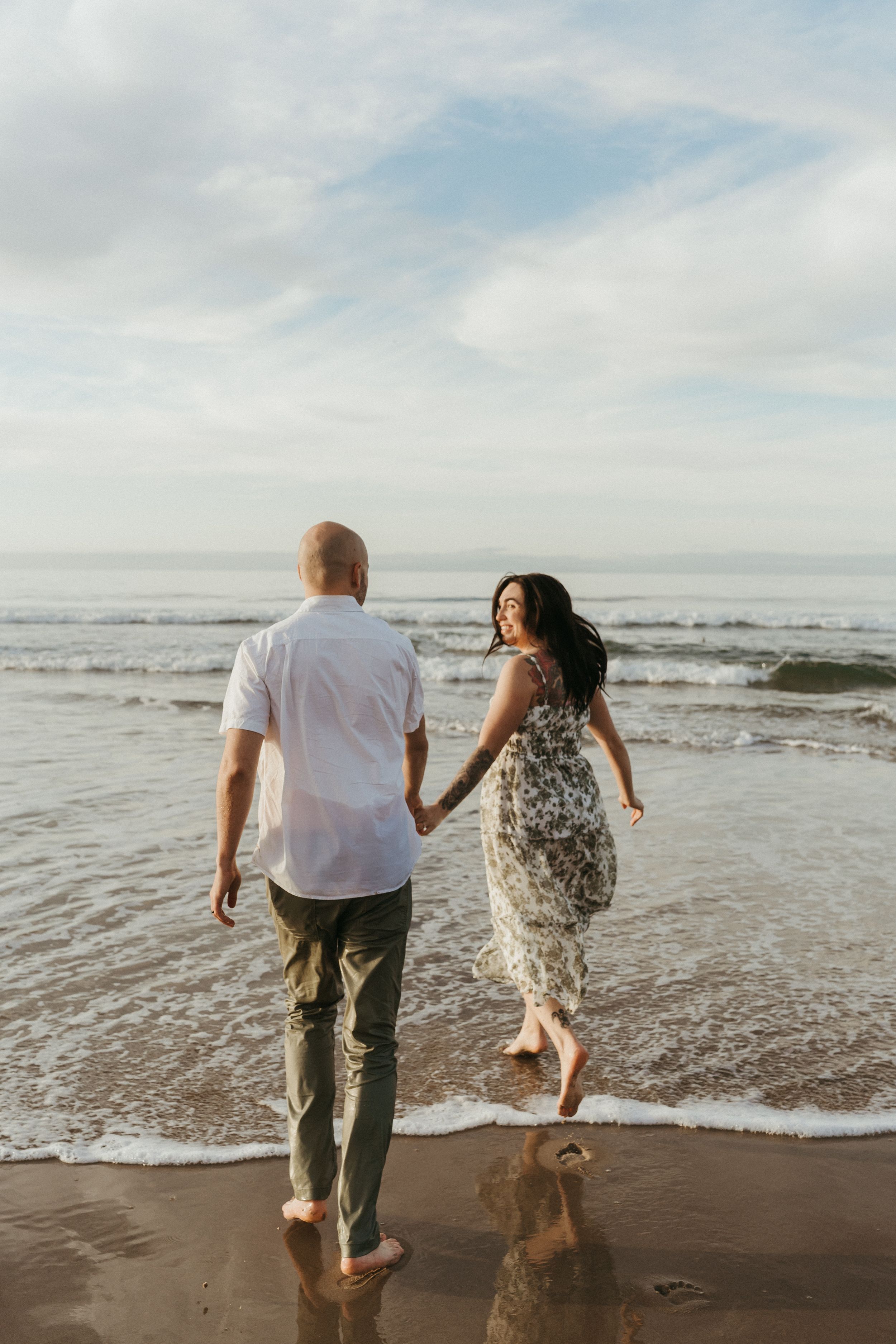 A romantic couple holds hands while walking barefoot along the beach shoreline at sunset.