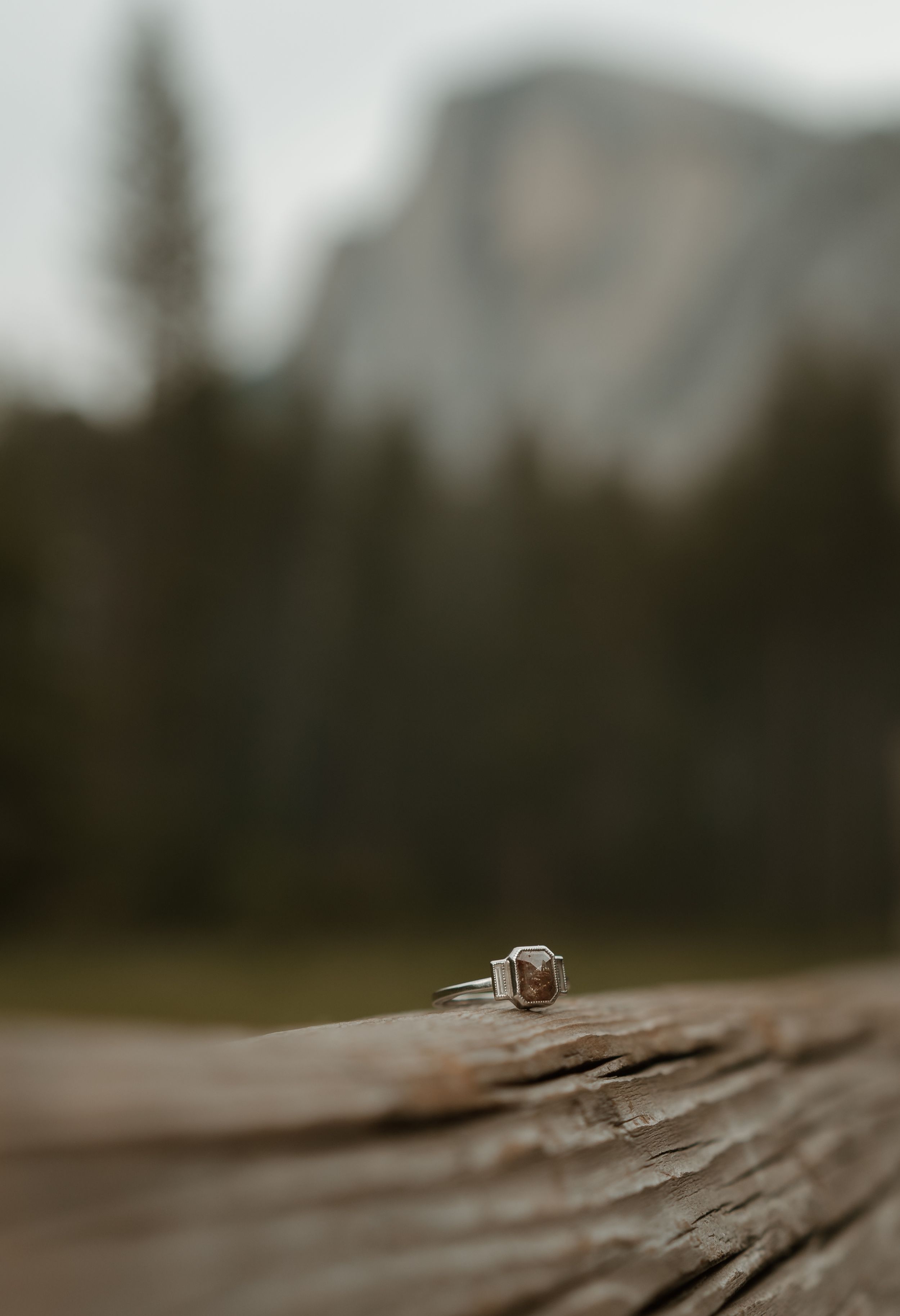A diamond engagement ring sits on weathered wood with Half Dome in Yosemite National Park blurred in background.