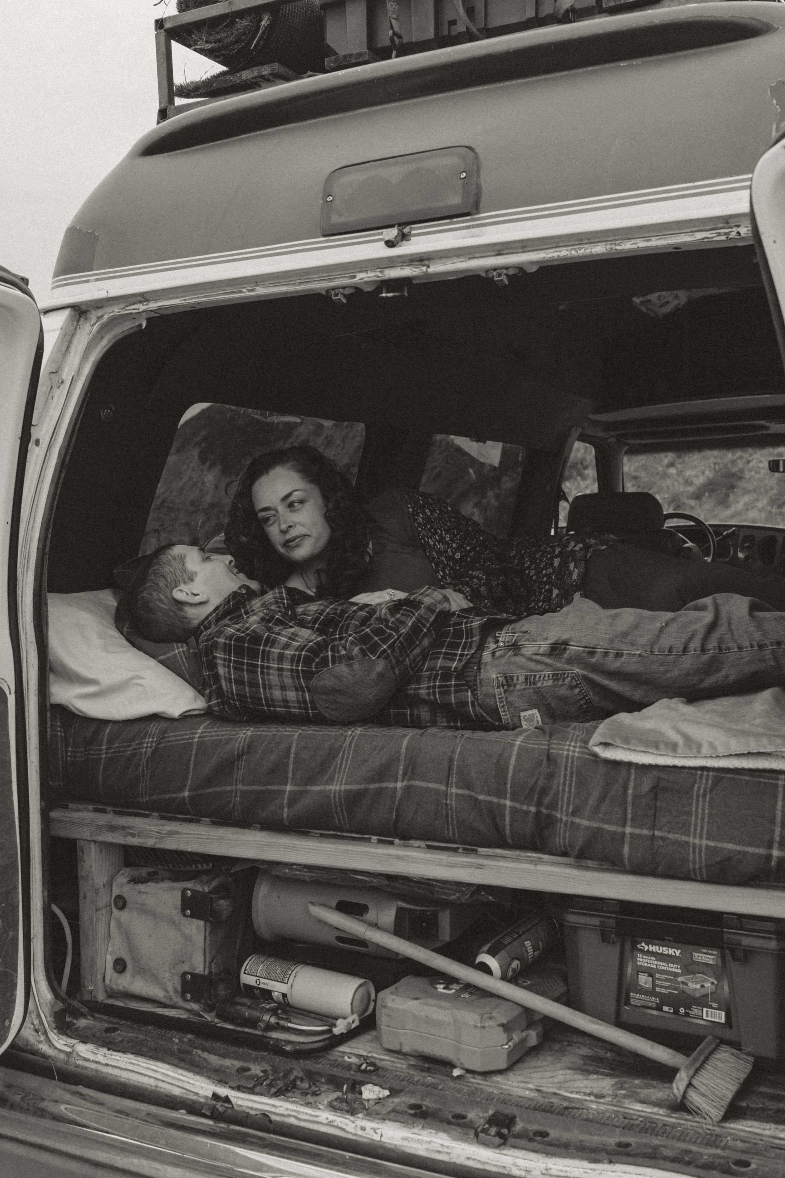 Black and white photograph showing people sleeping in the back of a van with plaid bedding and storage underneath.
