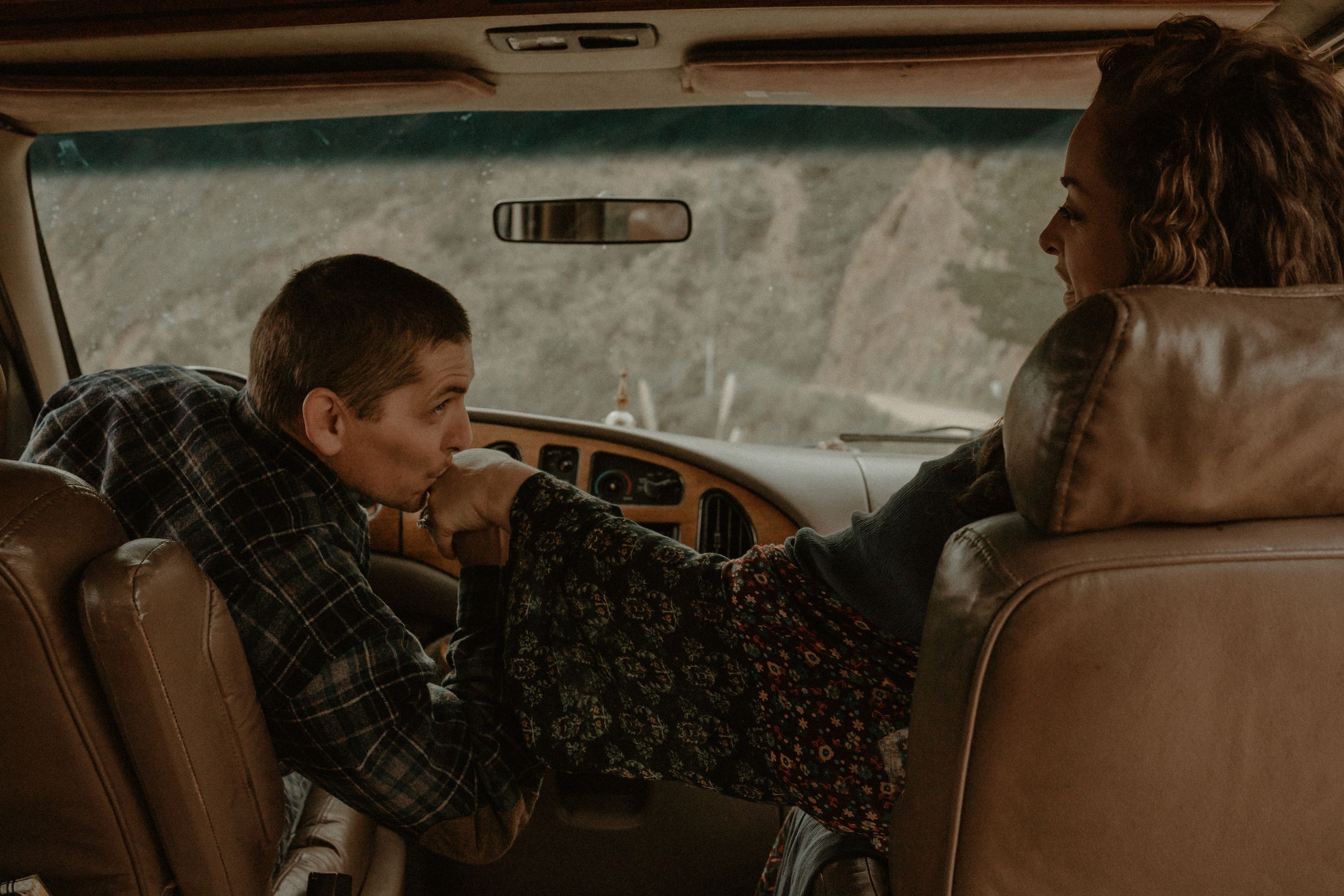 Two people sit in a vintage car with brown leather seats having a conversation in warm retro lighting.