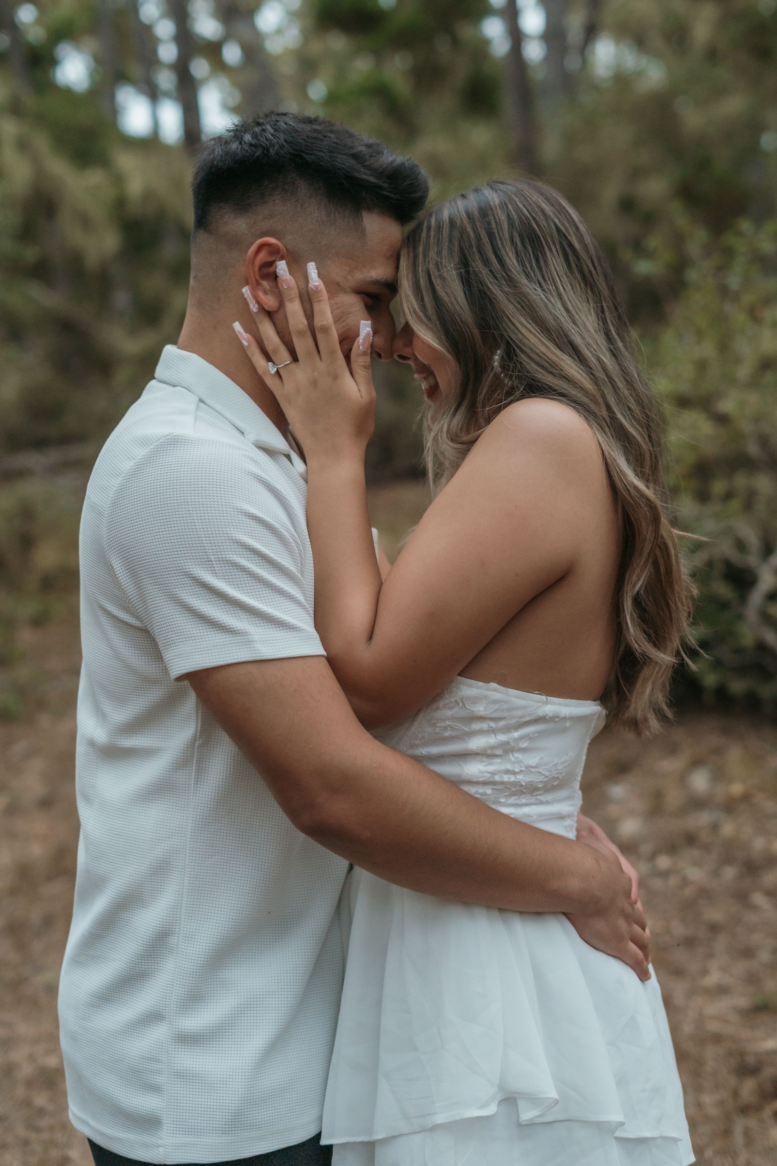 A romantic couple in white clothing share an intimate moment together on a wooded forest path.