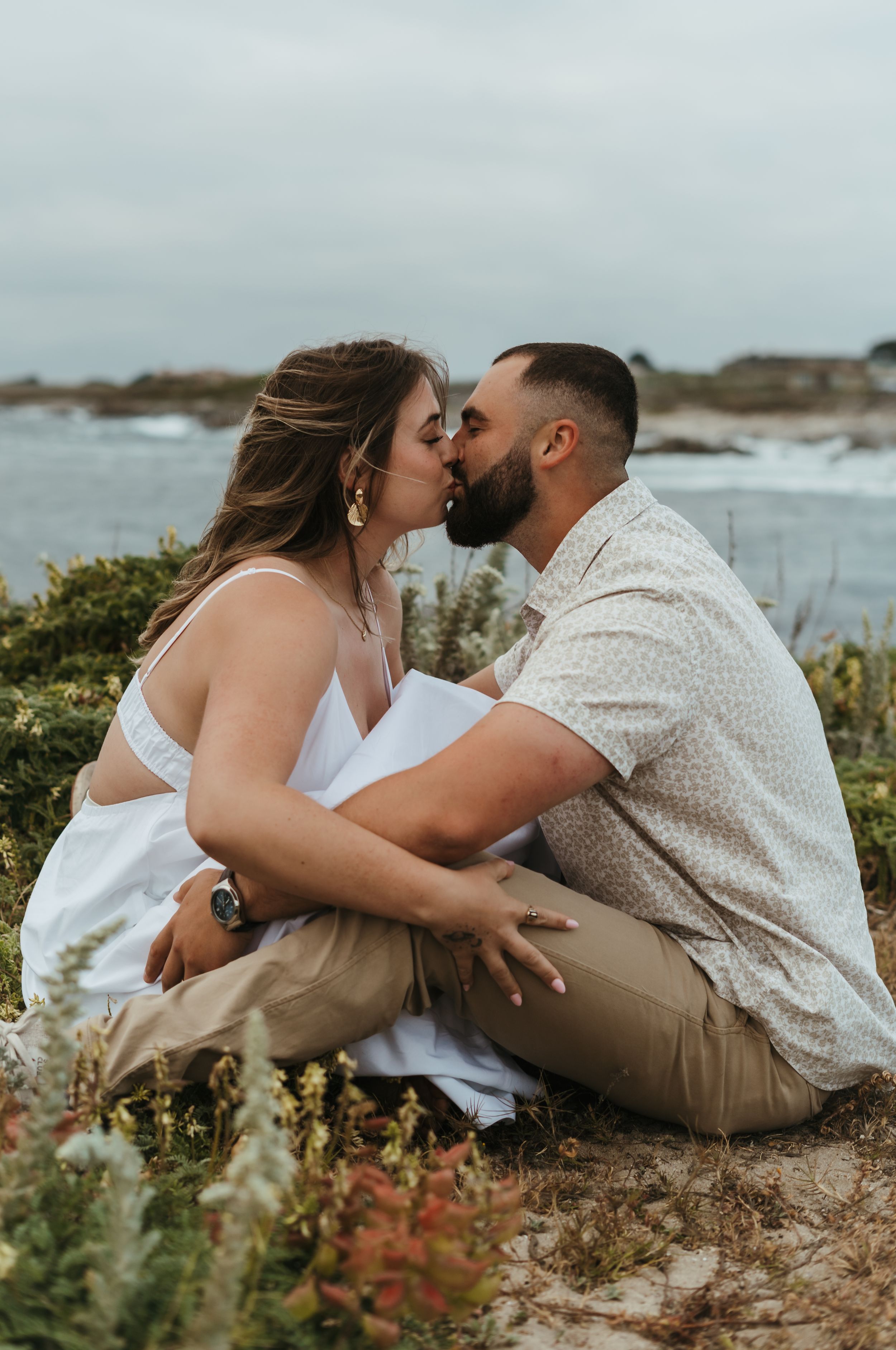 A romantic engagement photo shows a couple sharing an intimate kiss on a coastal cliff with ocean views in the background.