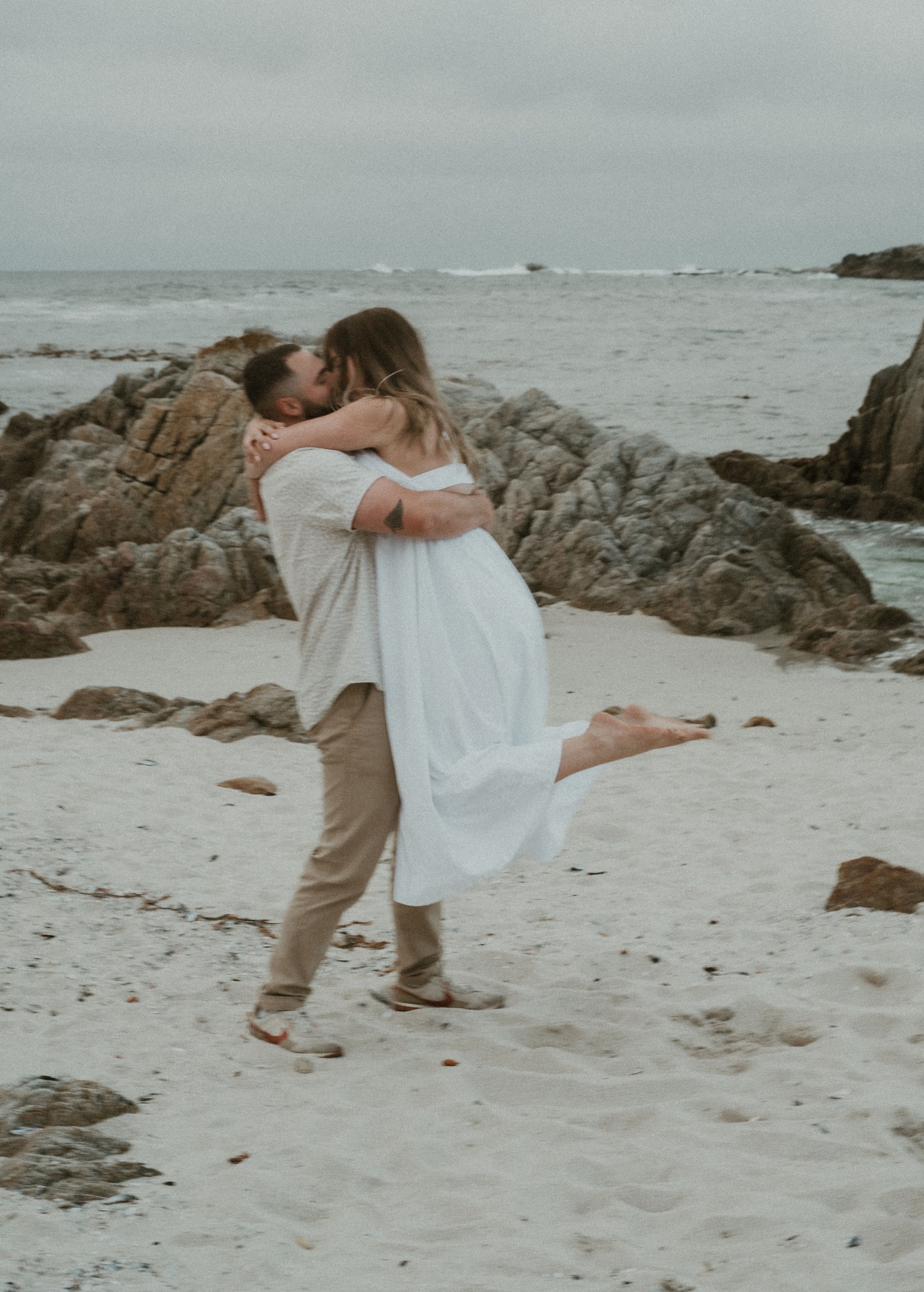 On a beach, two people dance together gracefully near rocky ocean shoreline during overcast day.