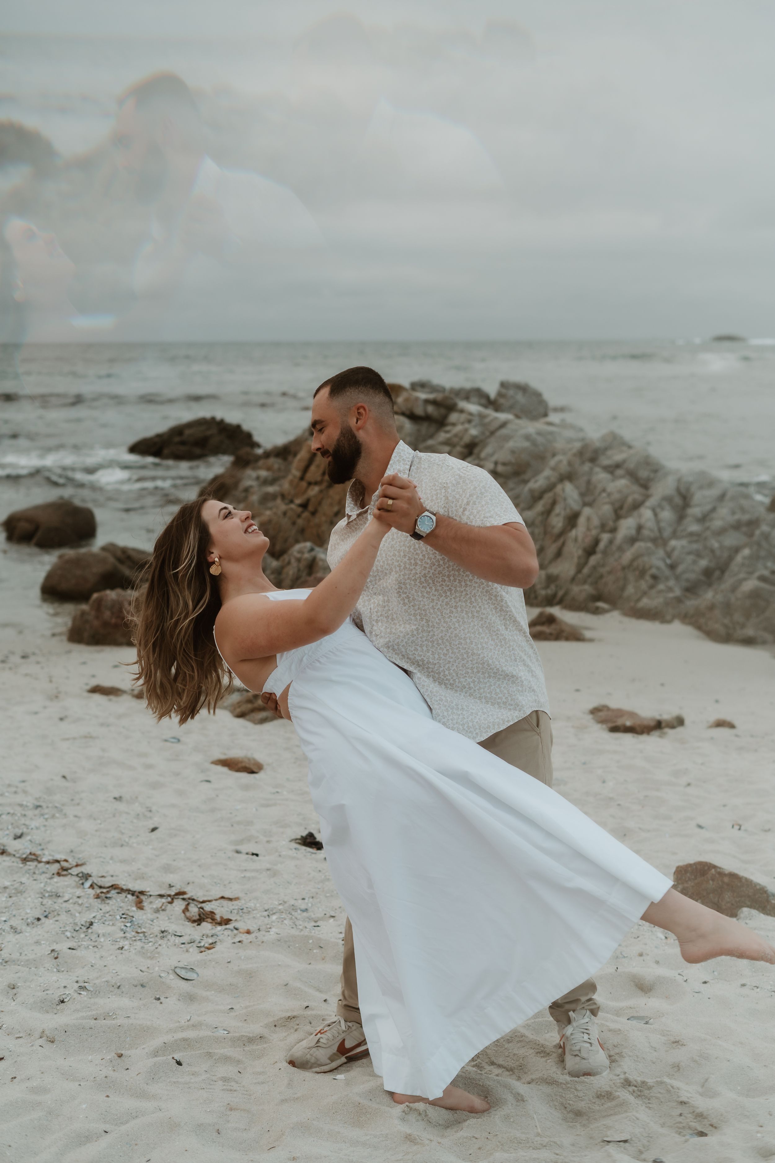 A couple in white outfits dances romantically together on a scenic beach with rocky outcrops near the ocean at sunset.