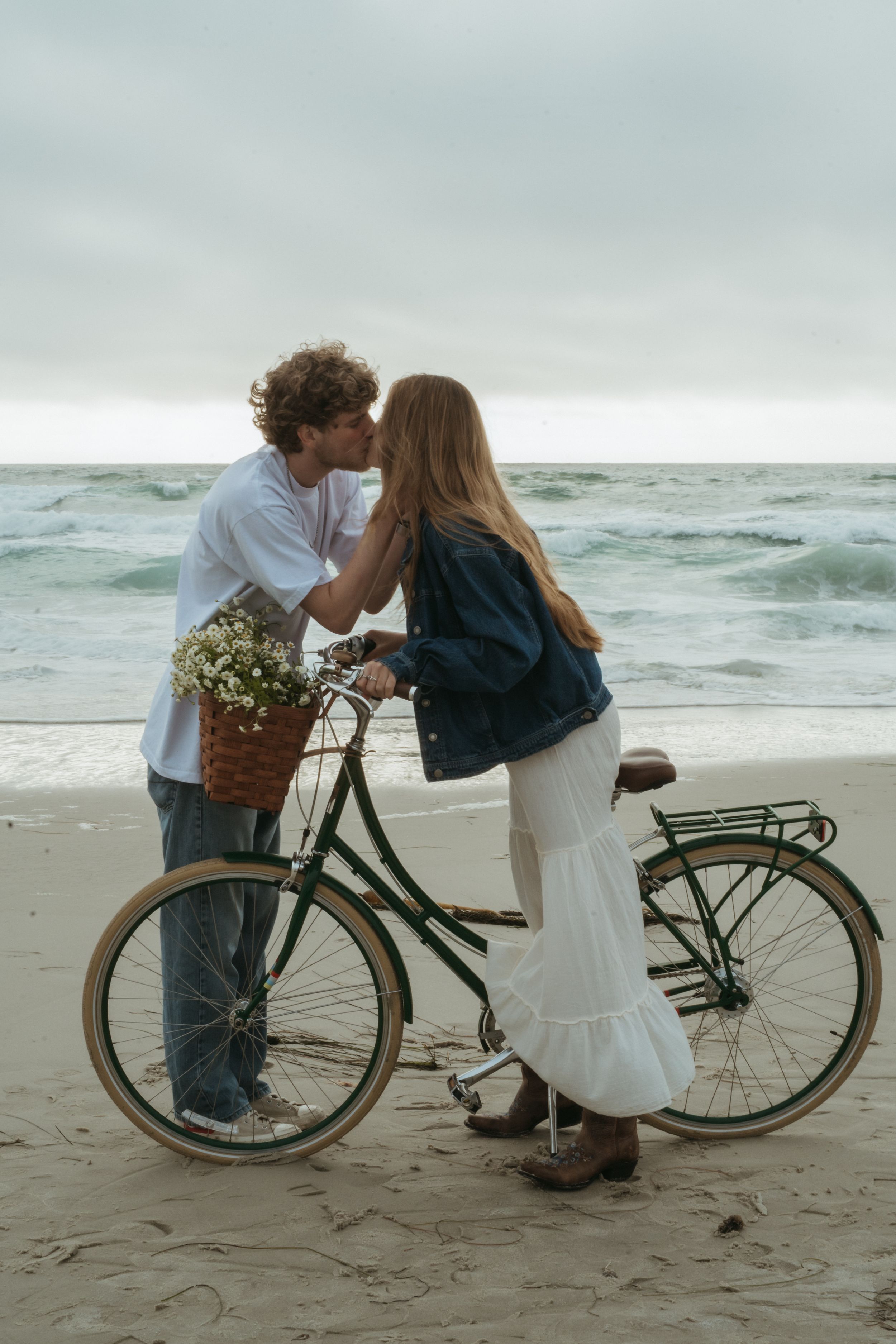 A romantic couple on a beach at dusk poses with a vintage bicycle adorned with flowers in the basket.